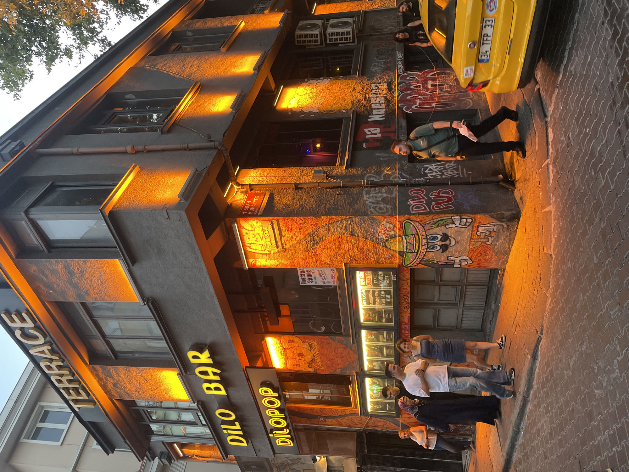 The image captures an exterior view of a building with a prominent sign reading "TERRACE" in large, illuminated letters. Below it, another sign displays "DILO BAR" in yellow, and adjacent to that, "DILOPPOP" is also illuminated. The building's facade is dark gray with dramatic orange lighting accentuating its architectural features. Graffiti adorns the lower portions of the building.
Several people are visible on the sidewalk. On the left, a group of three individuals stands near the building entrance. Further to the right, a couple walks along the street, the woman in a blue dress and the man in a white t-shirt and jeans. Another man in a green shirt walks past the building, carrying a white object. A bright yellow taxi with license plate "34 TFP 33" is parked on the right side of the street. The street is paved with cobblestones, and a brick sidewalk runs along the building. The scene appears to be in a public urban area, likely a commercial district. The overall ambiance suggests evening or nighttime, given the artificial lighting. This location is identified as Kadıköy, Turkey.