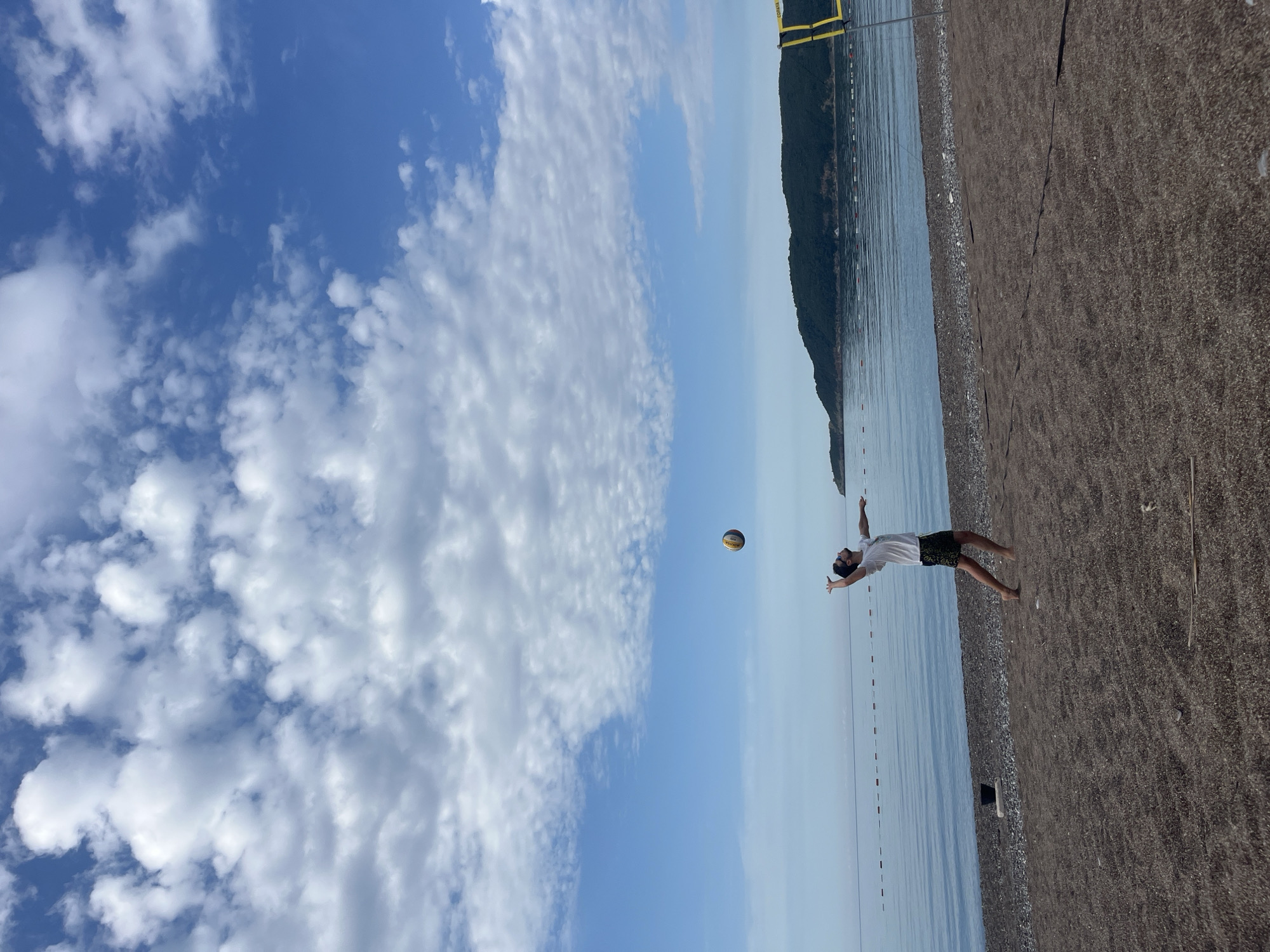 A sunny day at a beach in Boreti, Montenegro, showcases a man in a white t-shirt and patterned shorts leaping to hit a volleyball. The beach features dark sand, and the water is calm. A mountain range is visible across the bay. A yellow beach volleyball net pole stands nearby. The sky is bright blue with scattered white clouds.