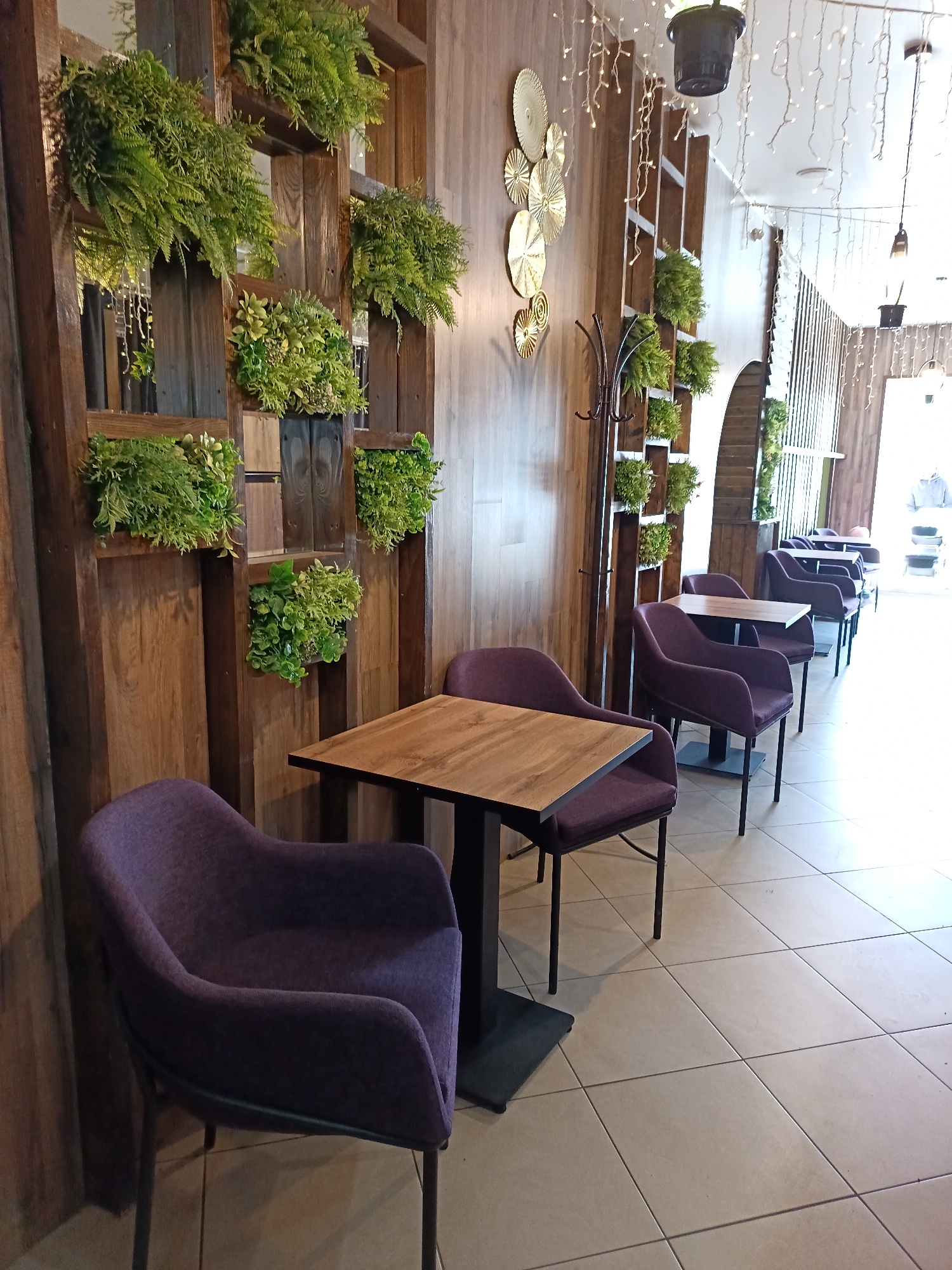 The image depicts the interior of a cafe or restaurant. The location is not stated in the prompt and therefore can't be determined. In the foreground, there is a table with a wooden top and a dark-colored base, surrounded by two purple upholstered chairs. The wall is adorned with a wooden structure featuring built-in plant holders filled with green foliage. Further into the space, there are additional tables and chairs of the same style, arranged in a row. A coat rack stands against the wall. Decorative elements such as metallic wall decorations and string lights are present. The floor is tiled in a light color. Natural light floods the space from an opening at the far end, creating a bright and airy atmosphere. There are no visible people in the scene.
