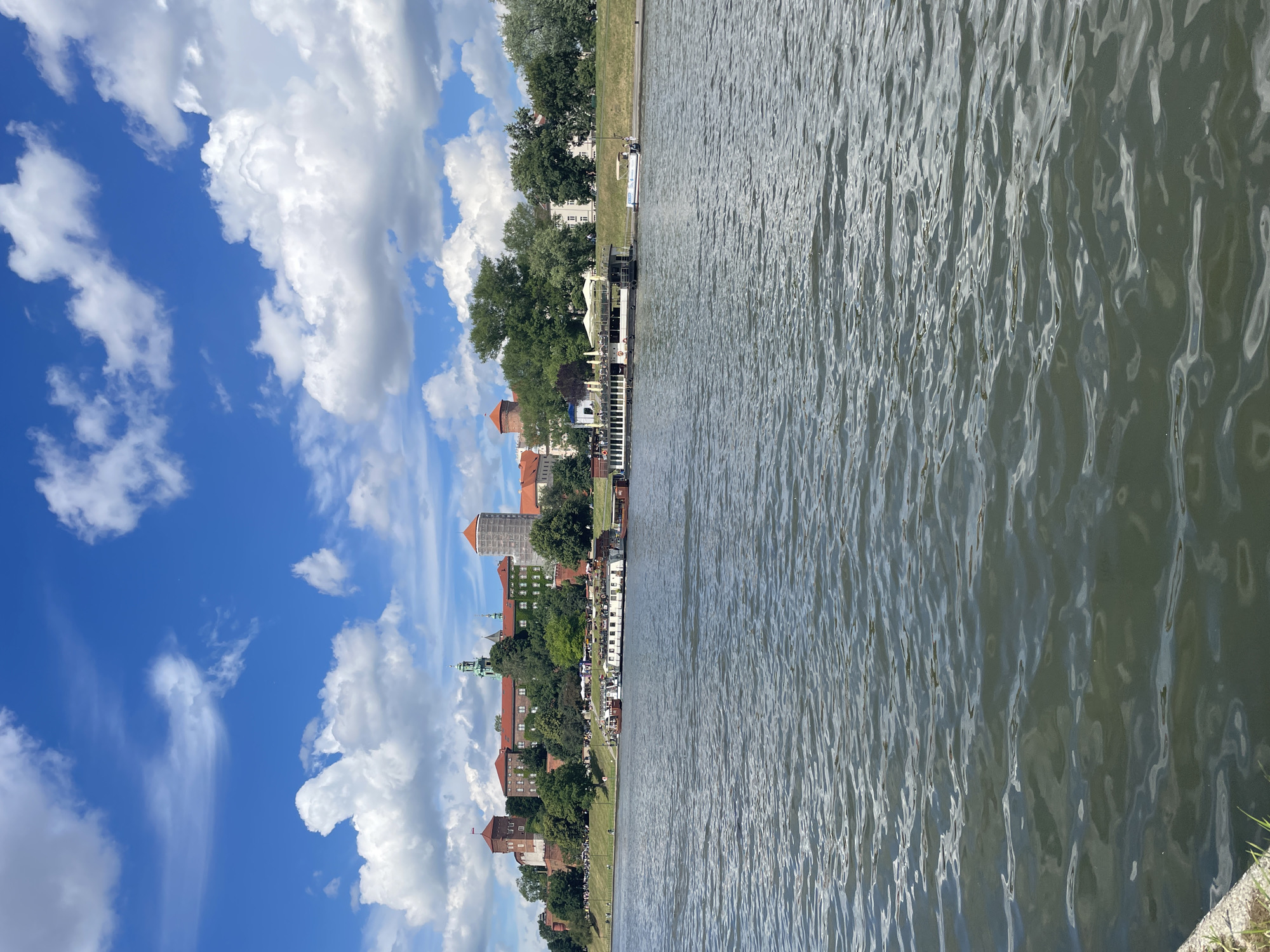 A wide, rippling river dominates the foreground of the image. In the background, a historic castle complex with red-tiled roofs and brick towers sits atop a tree-lined hill. Several boats are docked along the riverbank, some appearing to be restaurants or tour boats. The sky above is a vibrant blue, dotted with fluffy white clouds.