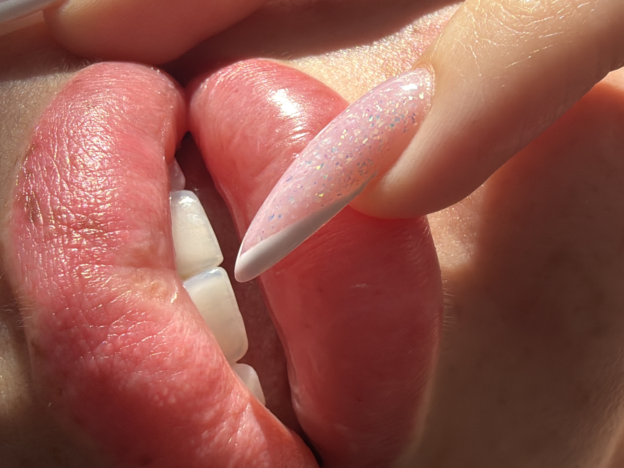 A close-up photograph depicts a person's lips slightly parted, revealing upper teeth. The lips are reddish, with visible texture. A manicured fingernail with a light pink base and white tip is positioned near the lips. The skin surrounding the nail is light-toned. There is no indication of location within Saint Petersburg, Russia, or any other city.