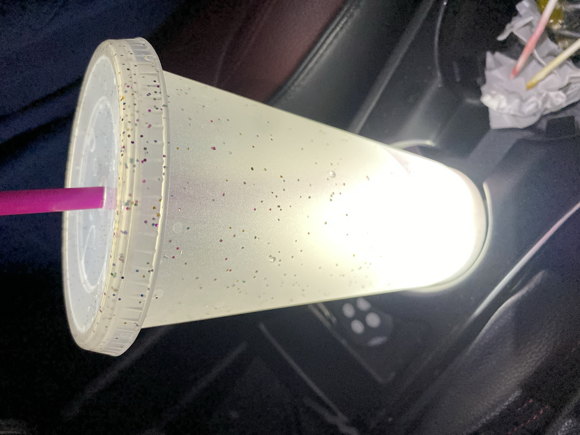 A plastic cup with a lid and straw sits inside a car. The cup is translucent with glitter, and a pink straw protrudes from the lid. Inside the cup, there is a light shining brightly. The cup is resting on a dark surface in the car. In the background, there is a car seat, and a tissue with what appears to be sticks. Puebla, Mexico is not depicted in the image.