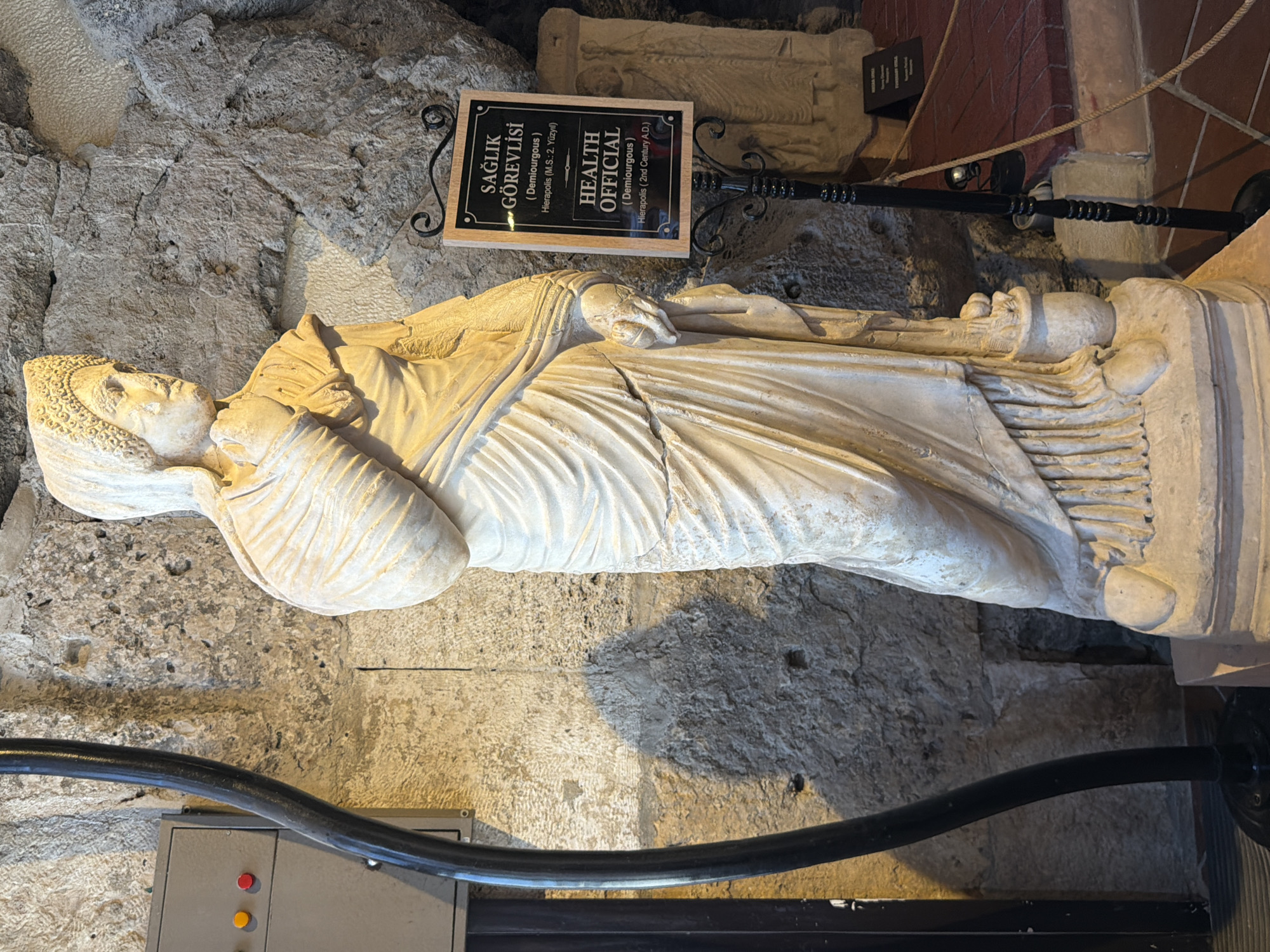 A full-body, standing marble statue of a woman is positioned in the foreground, facing to the right. The statue depicts a figure draped in flowing robes, with her left hand holding what appears to be a scroll or document. Her right hand is raised to her chin, with her fingers resting there. She wears a headdress that resembles a woven cap adorned with small beads or pearls. The statue stands on a tiered pedestal.
To the right of the statue, a black metal signpost supports a wooden plaque with black text. The plaque is divided into two sections. The top section, in Turkish, reads "SAĞLIK GÖREVLİSİ (Demiurgous Hierapolis (M.S.:2. Yüzyıl)". The bottom section, in English, reads "HEALTH OFFICIAL (Demiurgous) Hierapolis (2nd Century A.D.)".
Behind the statue and signpost, the environment consists of rough-hewn stone walls, characteristic of ancient architecture. A thick, black, curved pipe runs vertically along the left side of the image, in front of the stone wall. Further in the background, to the right of the main statue, another, smaller stone relief is visible, depicting two figures. The floor is paved with terracotta tiles. The lighting suggests an indoor museum setting, likely within the archaeological site of Hierapolis in Pamukkale, Turkey, given the inscription on the sign.