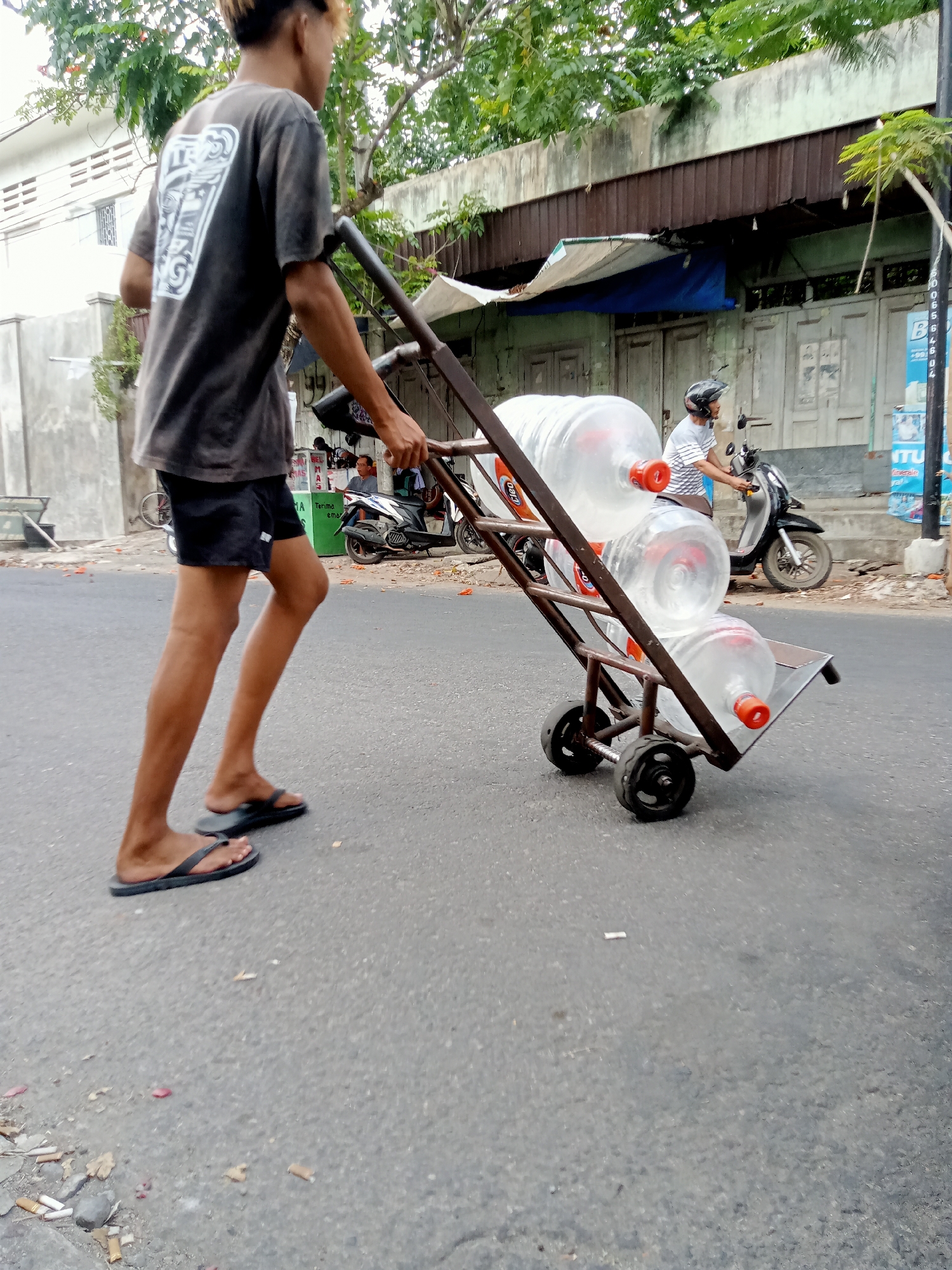 A young man with dyed hair walks down a paved street in Tulungagung, Indonesia, pushing a metal cart loaded with five large plastic water jugs. He is wearing a dark gray t-shirt with a graphic print on the back, black shorts, and black flip-flops. The cart has two wheels on the front and two smaller wheels in the back, and it appears to be made of rusted metal. The water jugs have orange caps and red labels.
In the background, a motorcycle with a rider wearing a helmet is parked on the side of the road. Behind the motorcycle, there is a building with wooden doors and a corrugated metal roof. Trees with green leaves line the street, and there are some scattered leaves and debris on the pavement. The overall scene appears to be a typical street in a residential or commercial area.