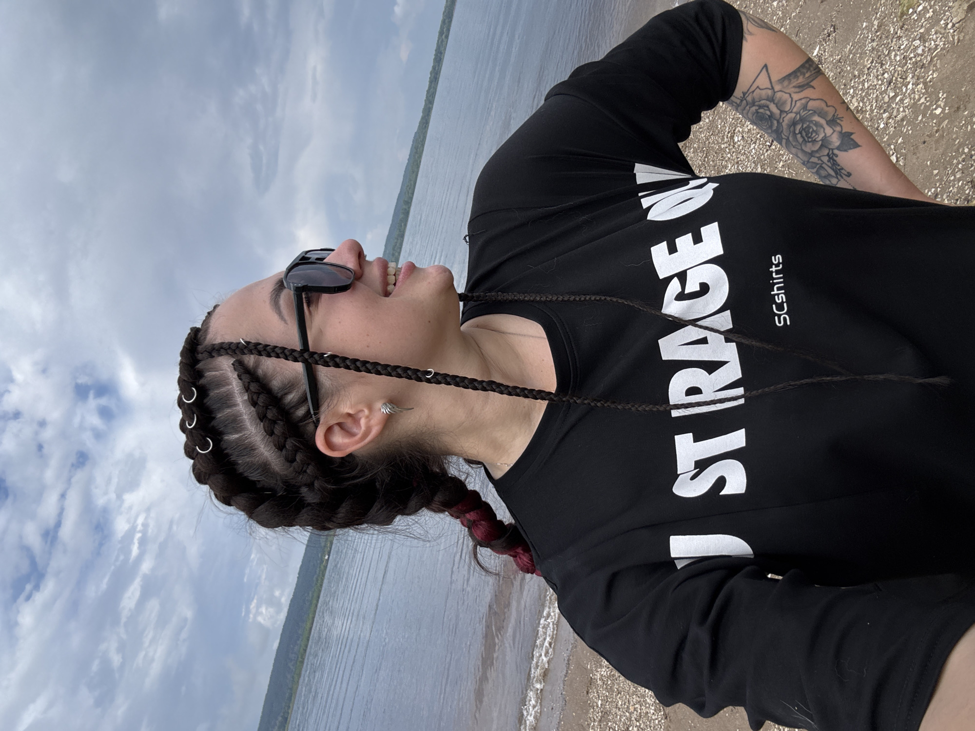 A woman with braided hair stands on a pebble beach near a body of water. She is wearing sunglasses and a black t-shirt with white text that reads "JUST RAGE ON" and "SCshirts". A tattoo of roses is visible on her right arm. The sky is cloudy.