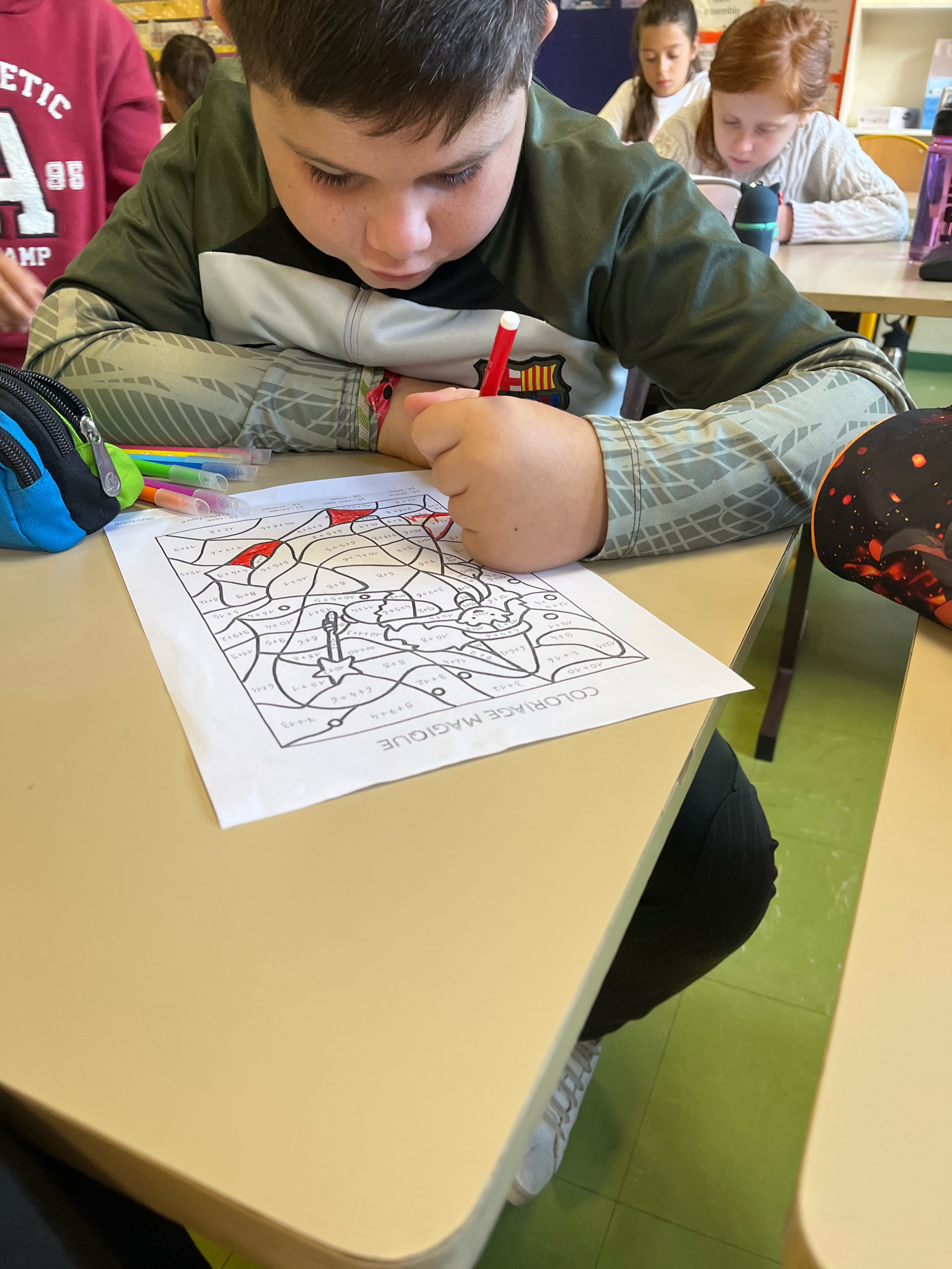 The image captures a classroom scene in La Fouillouse, France, focusing on a young boy engrossed in a math coloring activity. He is seated at a beige desk, his attention fixed on a worksheet filled with mathematical equations. A red marker is in his hand, poised to color in one of the sections. The worksheet itself features various mathematical problems, indicating a learning exercise.
In the foreground, a pencil case with blue and green accents rests on the desk, alongside a collection of colored markers and pencils. To the right, another pencil case with a dark background and fiery orange designs is visible.
In the background, two other children are also engaged with their schoolwork at adjacent desks. One child, a girl with long red hair, is wearing a cream-colored sweater and appears to be looking down at her paper. Another girl, with dark hair pulled back, is also looking down. The environment suggests a typical classroom setting with desks and possibly bookshelves visible in the distance. The floor is a vibrant green. The overall impression is one of focused learning.