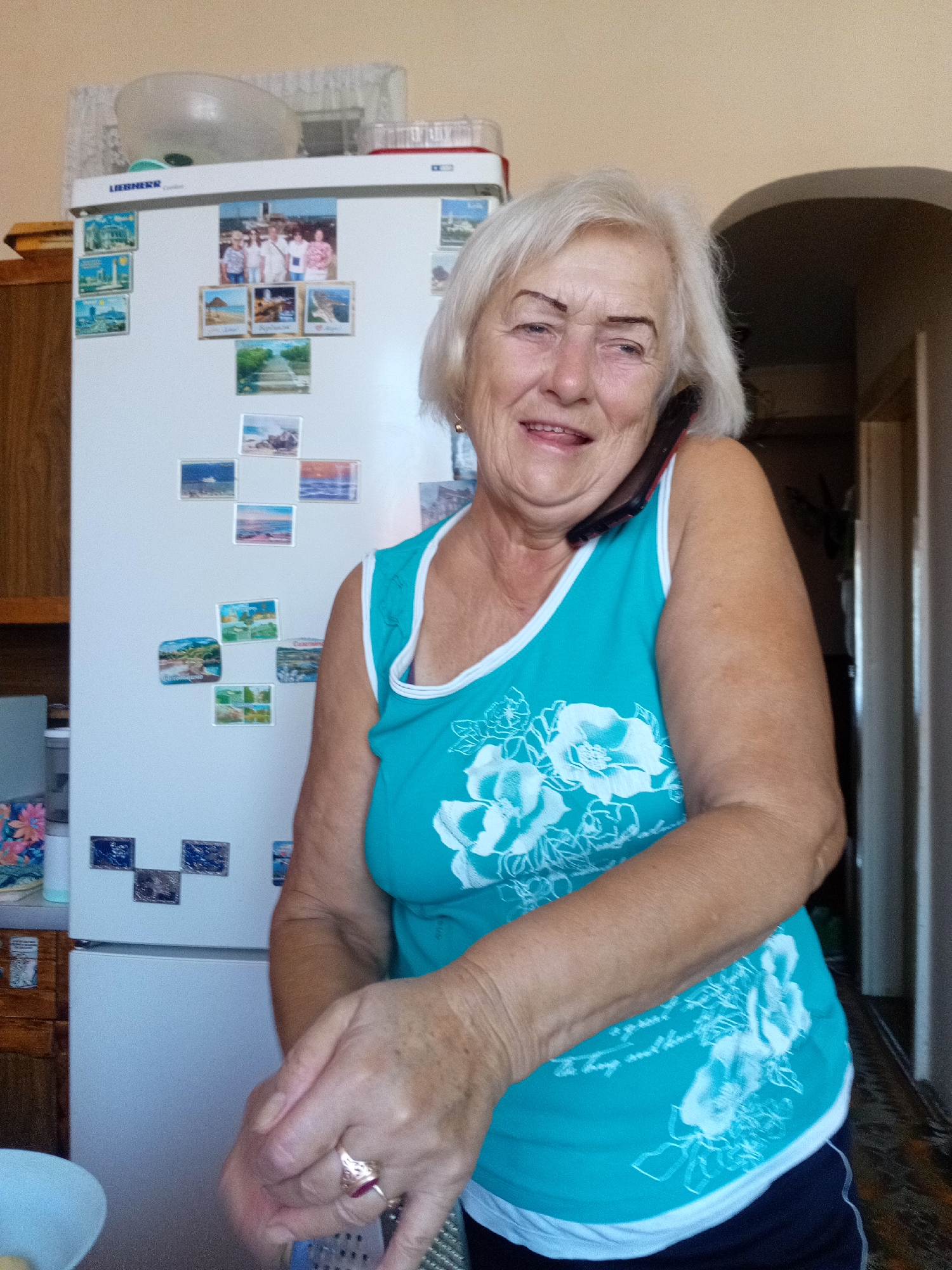 A medium shot captures an elderly woman with short, white hair, smiling while holding a dark-colored phone to her ear. She wears a turquoise tank top with a white trim and a floral pattern. She is standing in front of a white refrigerator, which is adorned with various small photographs, many depicting landscapes and family portraits. The woman's right hand is visible, extended downwards, and she appears to be holding a grater, suggesting she is engaged in food preparation. A ring with a red stone is on her ring finger. The background includes a wooden cabinet to the left and an arched doorway leading to a hallway on the right. The lighting suggests an indoor environment, possibly a kitchen. The scene is set in Tiachiv, Ukraine.