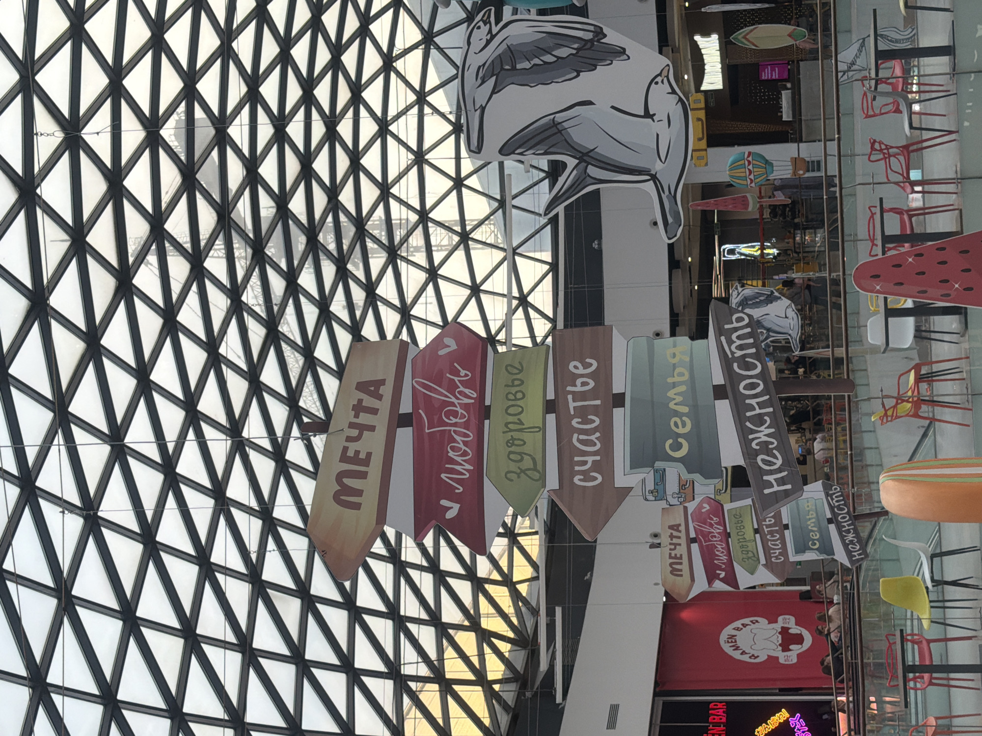 Here's a detailed description of the image:
**General Description:**
The image appears to be taken inside a commercial building in the city of Novoalexandrovka, Russia. The focus is on hanging decorative signage.
**Objects:**
* A large, geometrically patterned glass ceiling.
* Several signs are suspended from the ceiling. Each sign is shaped like an arrow and features a word written in Russian: "МЕЧТА" (Dream), "любовь" (Love), "здоровье" (Health), "счастье" (Happiness), "Семья" (Family), "нежность" (Tenderness).
* A large cartoon drawing of what seems to be a stork or seagull.
* A hanging hot air balloon.
* The signage for a "RAMEN BAR".
* Tables and chairs.
* A large watermelon.
**Environment:**
* The location appears to be a large indoor space, likely a shopping mall or commercial center.
* The area seems to be well-lit with natural light coming through the large glass ceiling.
**Additional Notes:**
The picture seems to be taken from below, looking up at the ceiling and the hanging decorations.