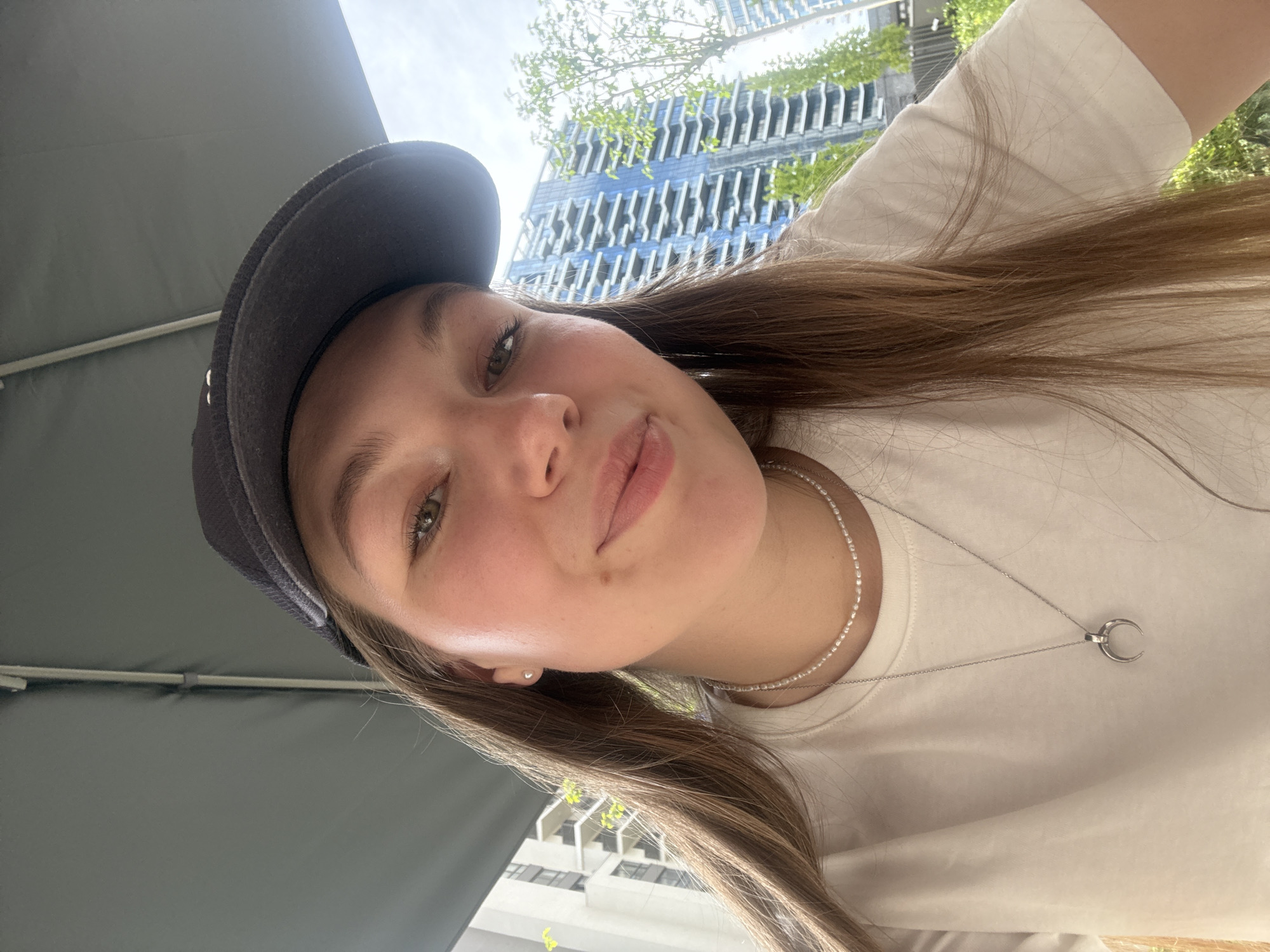 A young woman is taking a selfie outdoors, angled from below and slightly to the side. She is wearing a dark-colored baseball cap, a white t-shirt, and a layered necklace—one strand of small, round beads and another with a crescent moon pendant. Her long, light brown hair is parted and falls over her shoulder. The background features modern architecture and greenery. The buildings are identifiable as part of the Belgrade Waterfront development in Belgrade, Serbia. Part of an umbrella is also visible in the scene.