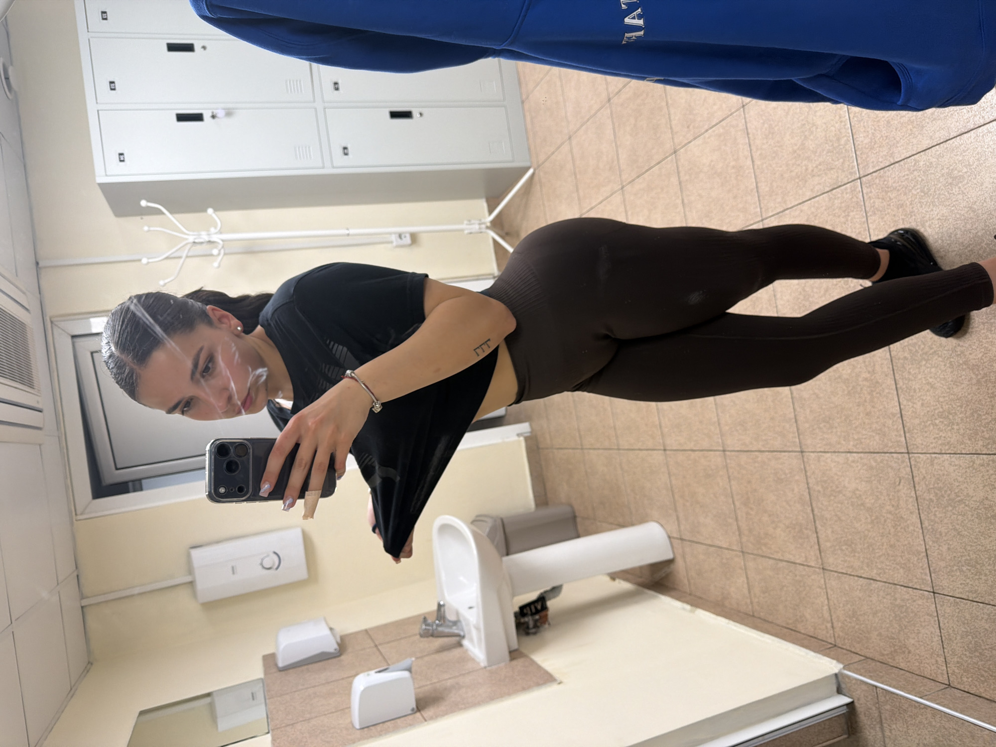 A woman is taking a selfie in a public restroom. She is holding a black iPhone in her right hand. The woman is wearing a black t-shirt and brown leggings with black shoes. She is standing in front of a mirror. The restroom has a sink, a paper towel dispenser, a soap dispenser, and a tiled floor. In the background, there are white lockers and a blue jacket or sweatshirt hanging up. The location in Gabrovo, Bulgaria, cannot be determined from the image.