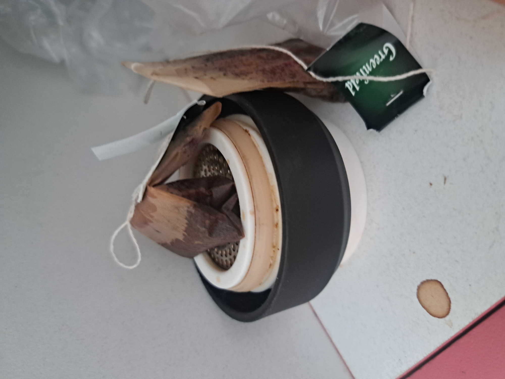 The image shows a top-down view of a tea infuser with a black outer ring and a white inner component, filled with used tea bags. The tea bags are brown and appear damp. A dark green tea tag with the word "Greenfield" is visible. The infuser sits on a surface that appears to be white with a brown stain near the bottom right corner. There is also a plastic bag partially visible in the upper left of the image. The photograph appears to have been taken in a low-light environment. There is no evidence in the photo to confirm that it was taken in Leningradskaya, Russia.
