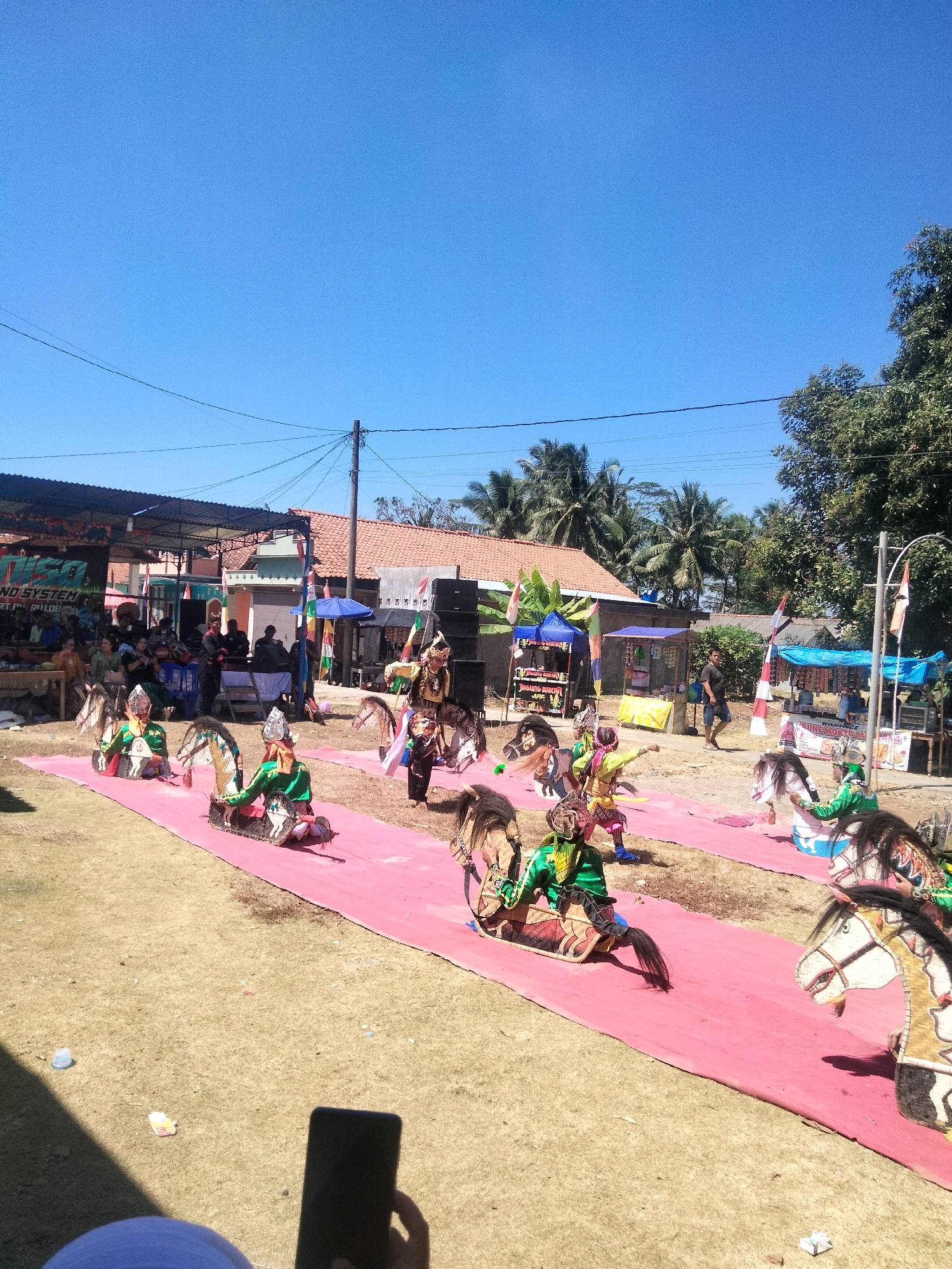 The image depicts a traditional cultural performance, possibly "Kuda Lumping," taking place outdoors in Cilacap, Indonesia. Several performers are dressed in colorful costumes, some wearing headdresses and elaborately decorated horse effigies around their waists. They are positioned on a pink carpet laid out on a grassy area. In the background, there are temporary stalls or tents, a stage with sound equipment, and spectators gathered. Palm trees and other vegetation are visible, suggesting a tropical or subtropical environment. The sky is clear and blue, indicating fair weather. The overall scene captures a lively community event.