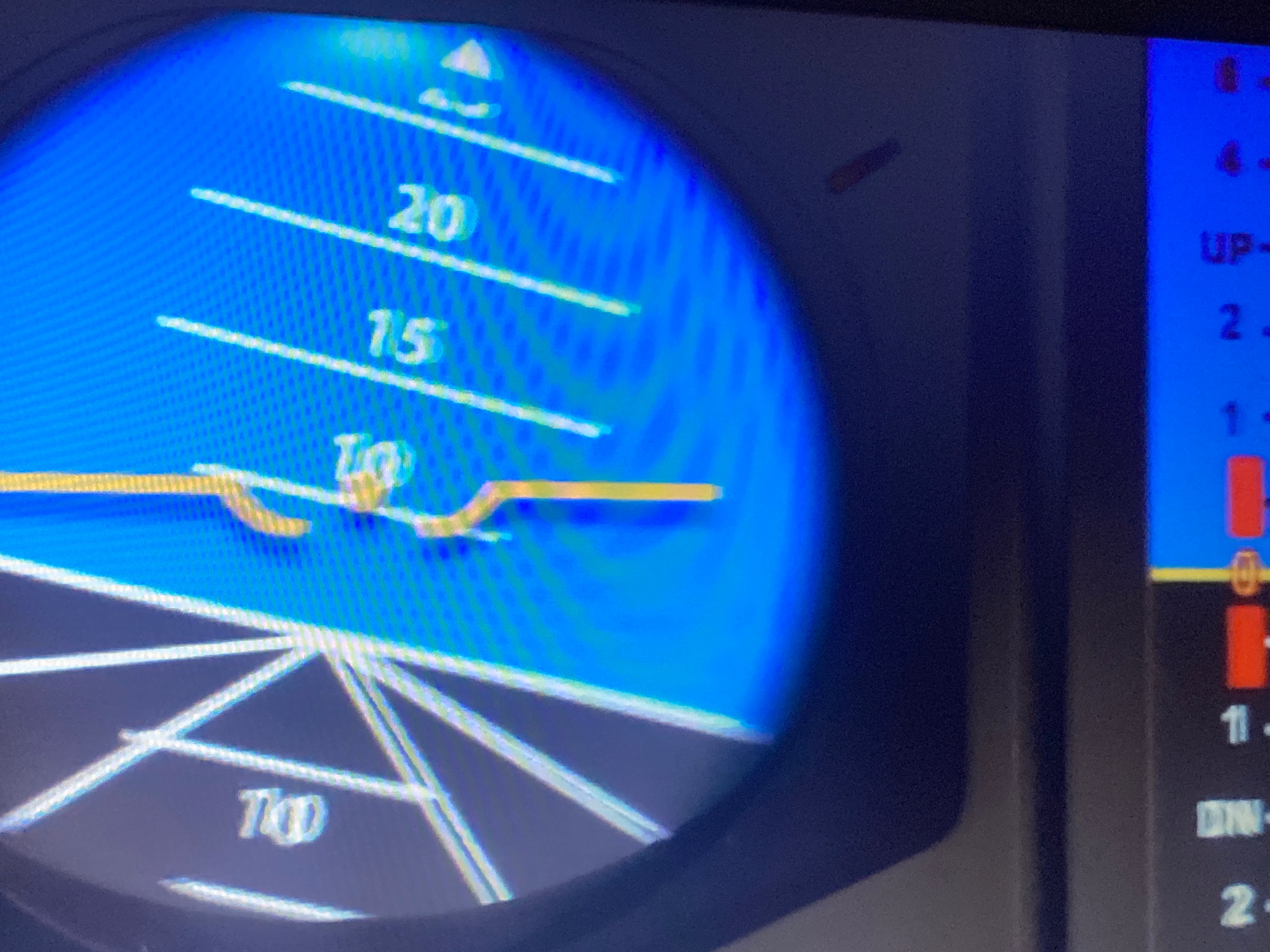 The image shows a close-up view of an aircraft instrument panel, likely a digital Artificial Horizon or Attitude Indicator, against a dark background. The primary display is a bright blue screen with white markings and digital readouts. On the left side, a circular display depicts a horizon line with an aircraft symbol above it. Markings for altitude or vertical speed are visible, with numbers like "20", "15", and "10" displayed. A yellow bar likely indicates the aircraft's current pitch or attitude. On the right side, a vertical blue display shows numerical data with red and white bars, possibly indicating system status or further flight parameters. The lighting suggests an indoor environment or a dimly lit cockpit. The image appears to be taken from a slightly upward angle. There is no discernible indication of the location being in Hemayetpur, Bangladesh, or any specific city.