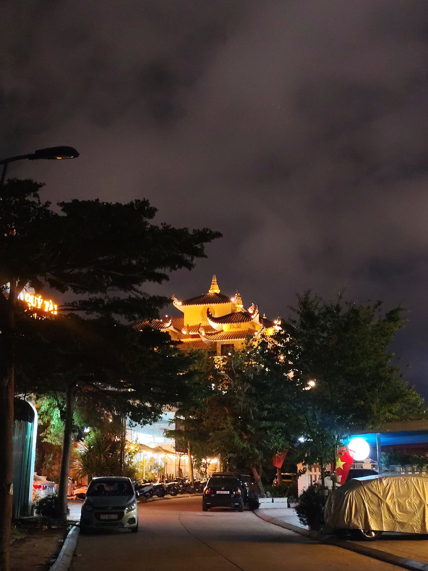 The image captures a night street scene in Da Lat, Vietnam. A curved road leads towards a brightly illuminated pagoda-style building, the focal point of the scene. The building's multiple tiers and ornate roofline are distinctly visible against the dark sky.
Several vehicles are parked along the roadside, including a light-colored compact car in the foreground and a black sedan further down the road. A car covered with a reflective material is parked on the right side of the road.
Streetlights and the pagoda's illumination provide the main light sources, casting shadows and highlighting the surrounding trees and foliage. A street lamp is on the left side. The sky is dark, with areas of visible cloud cover.