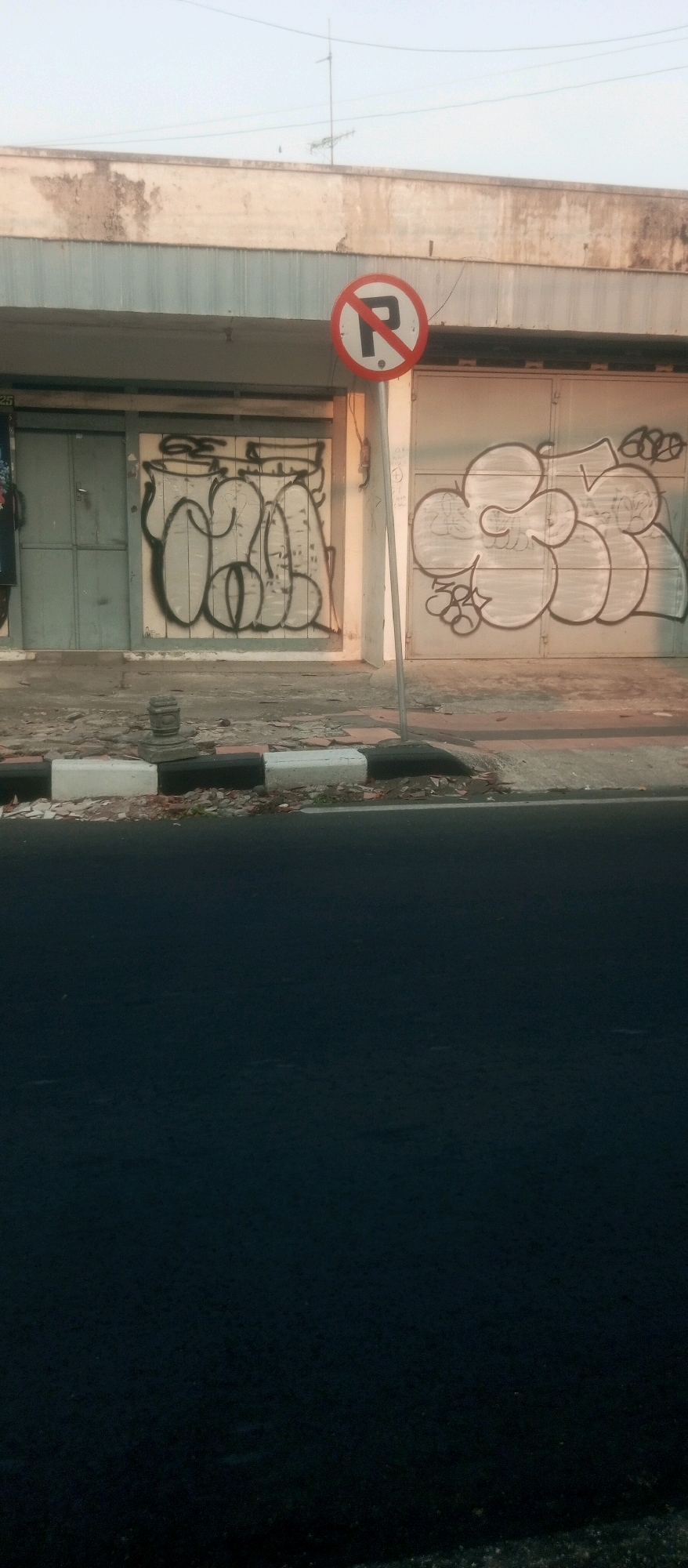 A "no parking" sign is affixed to a metal pole in front of a building with corrugated metal doors that have graffiti art. The building is on a street in Kediri City, Indonesia. The street has a curb with a pattern of black and white blocks. The road surface is asphalt. The sky is clear and bright.