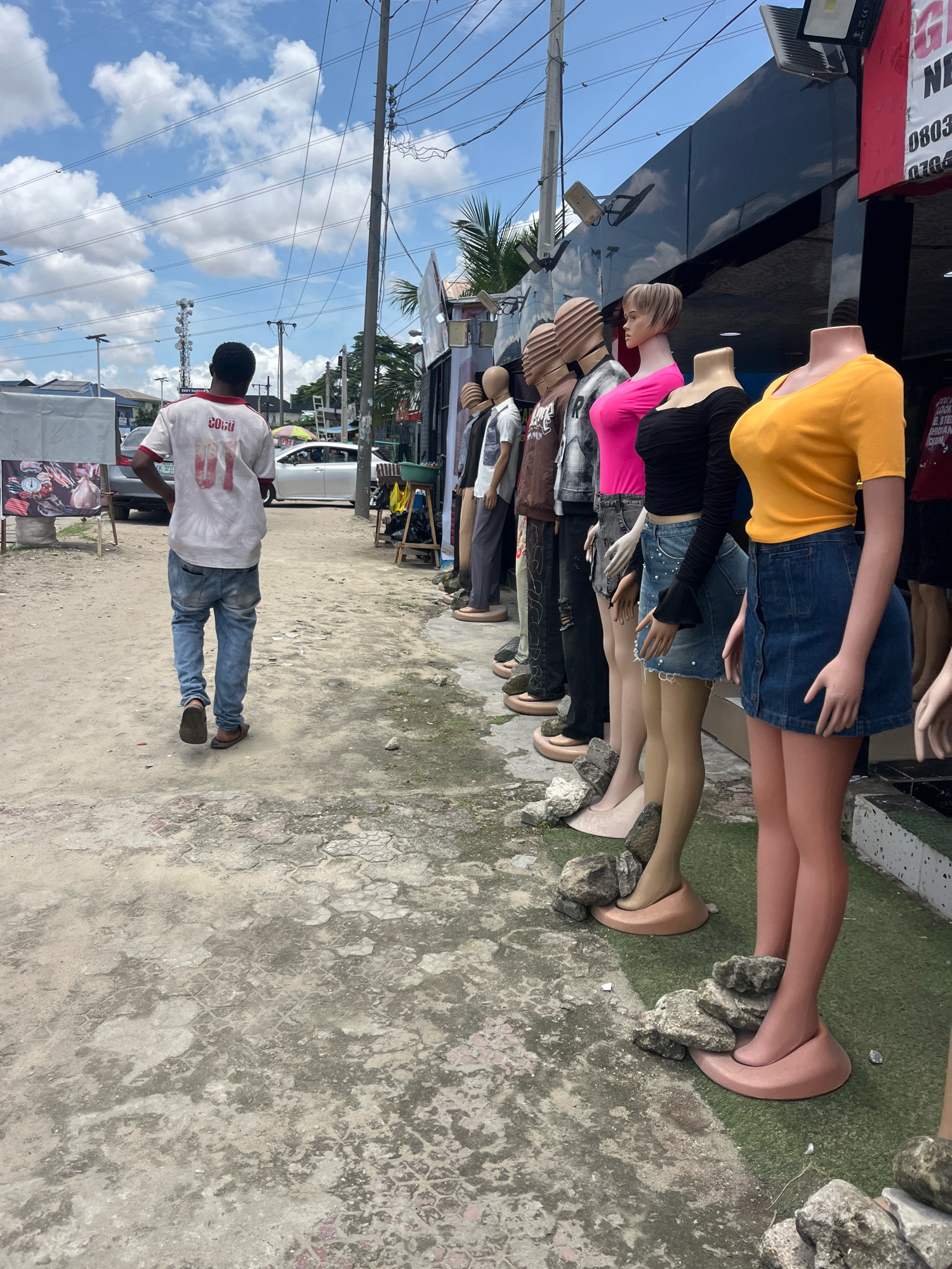 In Ado, Nigeria, a person walks away from the camera on a concrete path. The person is wearing a white t-shirt with red lettering and blue jeans. To the right of the path, a row of mannequins is displayed outdoors in front of a storefront. The mannequins are dressed in various clothing items, including a yellow t-shirt, a black long-sleeved shirt, a pink t-shirt, and other outfits. Some mannequins are missing heads. The storefront has a dark facade with lights mounted on poles and a red sign with white lettering. Overhead, power lines and clouds are visible against a bright sky. The environment appears to be a commercial area with some vehicles and other structures in the background.