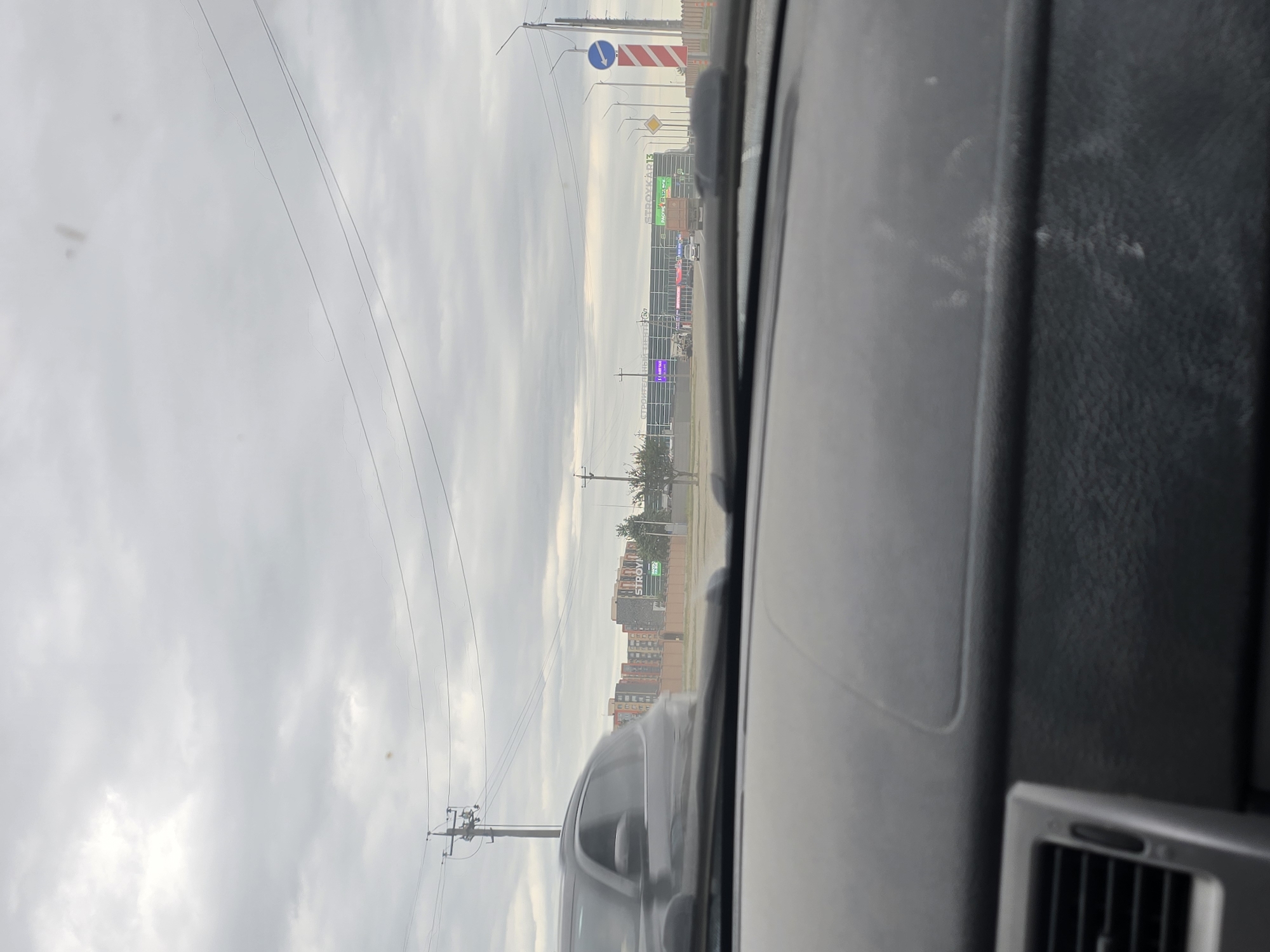 The image depicts a view from inside a vehicle, looking out through the windshield. The scene is set in the city of Grozny, Russia. In the foreground, the dashboard of the car is visible, with a vent on the left side. Beyond the windshield, a street scene unfolds under a cloudy sky.
On the right side of the road, a large commercial building is visible with signage that appears to read "СТРОЙКАРК13" (STROYARK13). Adjacent to it, another sign reads "СТРОИТЕЛЬНЫЙ ЦЕНТР" (BUILDING CENTER). Traffic signs are also present, including a blue directional sign with a white arrow pointing left and a red and white striped pole.
Several vehicles are on the road, including a dark-colored SUV in the left middle ground, moving past the camera. A truck and other cars are visible further down the road. In the background, residential buildings with multiple stories and a distinctive architectural style are visible. Utility poles with power lines crisscross the sky. The overall atmosphere suggests a suburban or commercial area within the city.