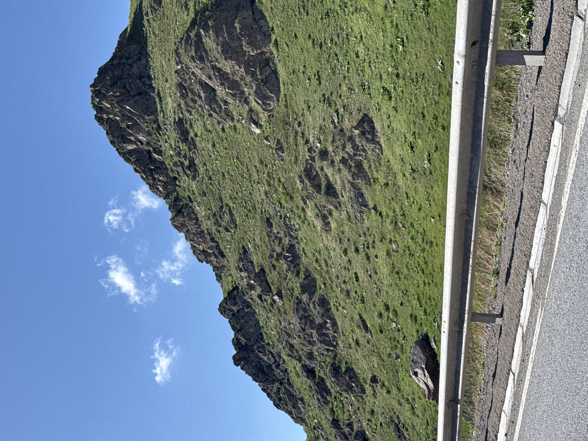 A landscape scene showing a grassy, rocky mountain slope next to a road. The road has a metal guardrail along its edge and a white stripe marking the lane. The mountain is covered in green vegetation. A blue sky with small clouds is visible on the left side of the image. The photograph appears to be taken from a slightly low angle, looking up at the mountain. The location is not verifiable.