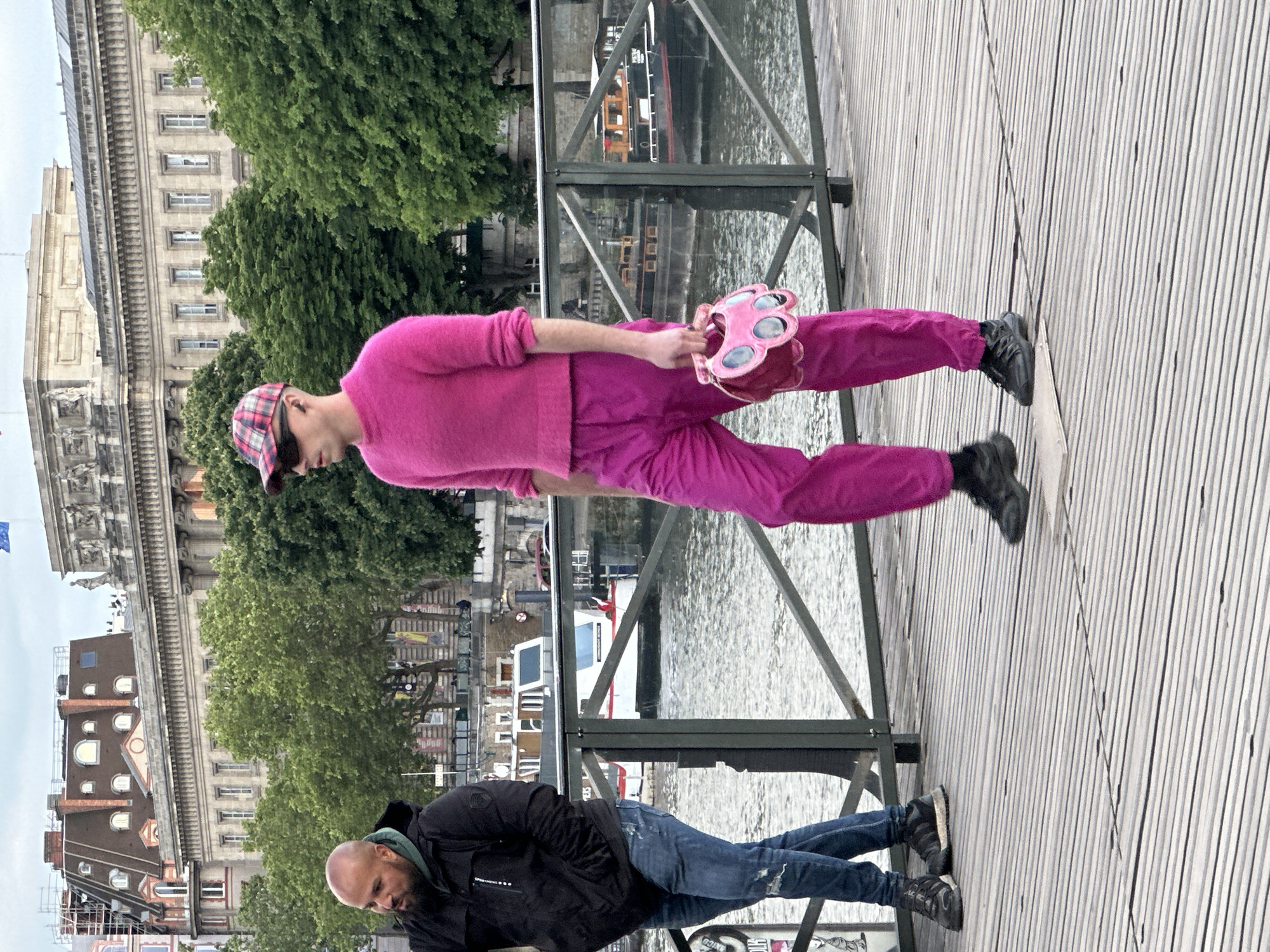 A full shot shows two men on a bridge in Paris, France, with buildings, trees, and a river visible in the background.
One man is wearing a bright pink outfit including a sweater, pants, and a plaid baseball cap. He is also holding a pink purse that looks like an animal paw. He is standing and facing towards the river.
The other man is wearing a black jacket and jeans. He is looking down.
The bridge has a wooden surface and glass railings.