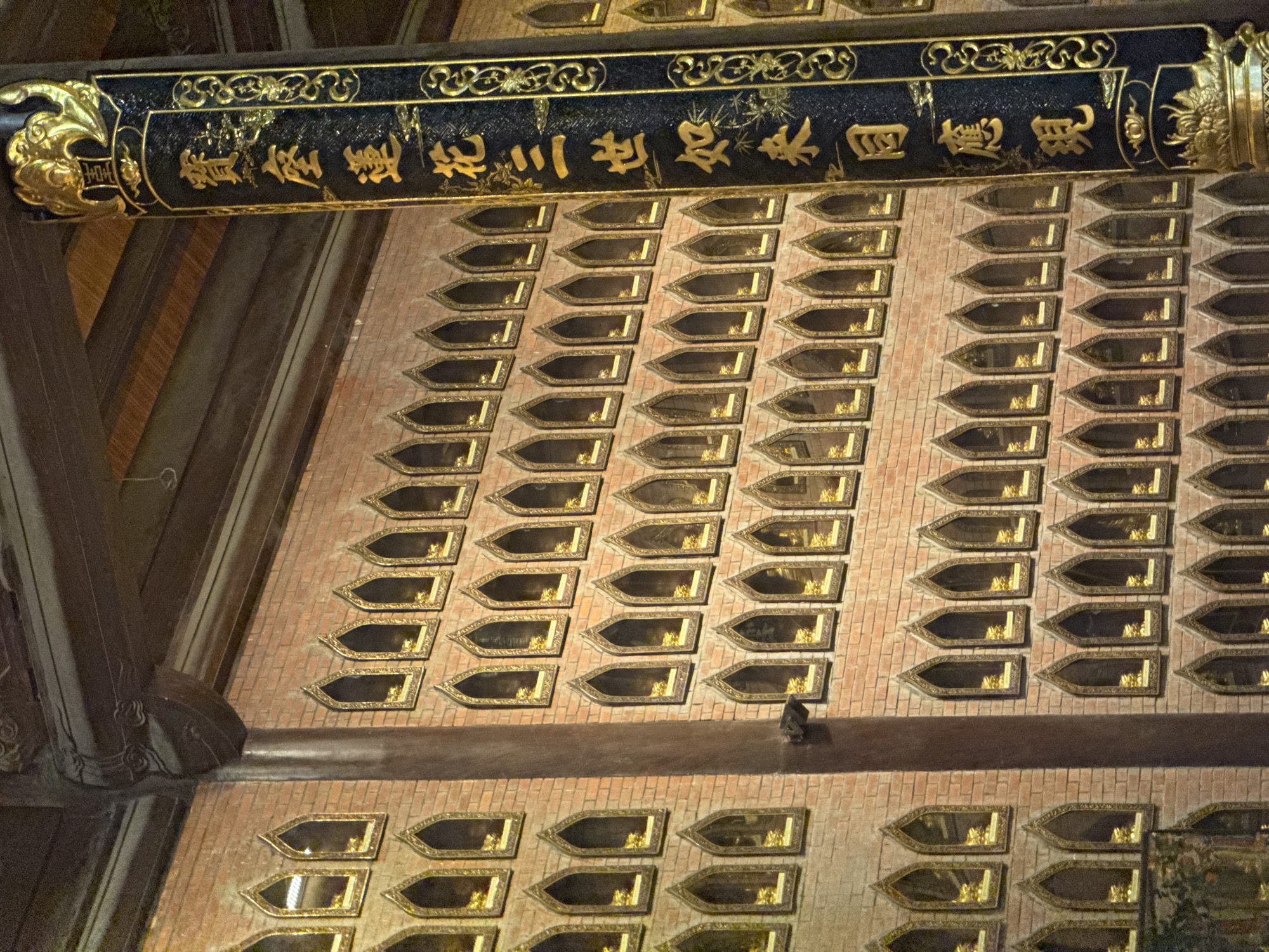 The image shows a brick wall lined with hundreds of small, arched niches, each containing a small golden statue, likely of Buddha. The niches are framed with gold trim and appear to be set into the brickwork. To the right of the wall is a large, dark pillar adorned with gold calligraphy and decorative elements. The ceiling above is visible in the upper portion of the image, appearing to be made of wood. This scene is located within the city of Tây Hoa Lư Ward, Vietnam, and is likely part of a Buddhist temple or sanctuary. The overall impression is one of ornate religious architecture and devotion.