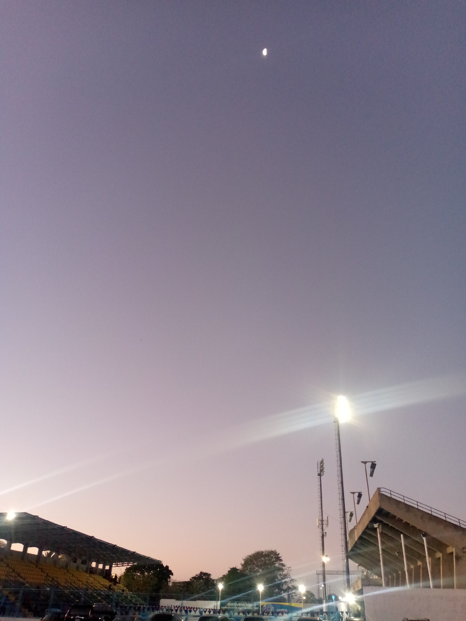 The image depicts a stadium setting at dusk or dawn in Unknown, Unknown. A crescent moon is visible in the upper portion of the sky. Below, portions of stadium seating are seen, along with tall light poles illuminating the scene. Cars and festive banners are visible near the base of the stadium.