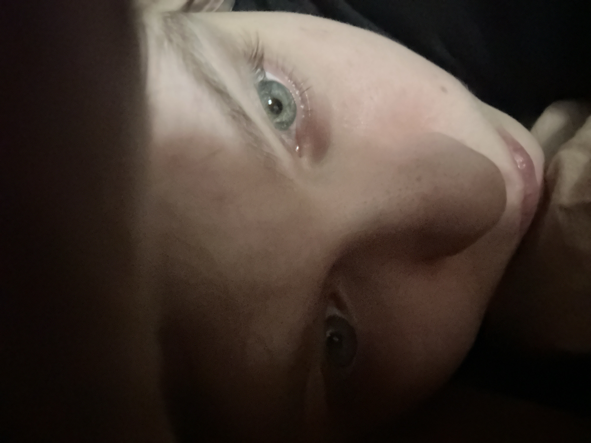 A close-up, low-angle shot focuses on a person's face, featuring an eye with light-colored iris and a single tear. The nose is prominent and the lips slightly parted. The skin tone is pale, with visible pores on the nose. The background is dark, obscuring details but suggesting clothing or another dark surface. The photograph does not contain enough information to discern a location, including Moscow, Russia.