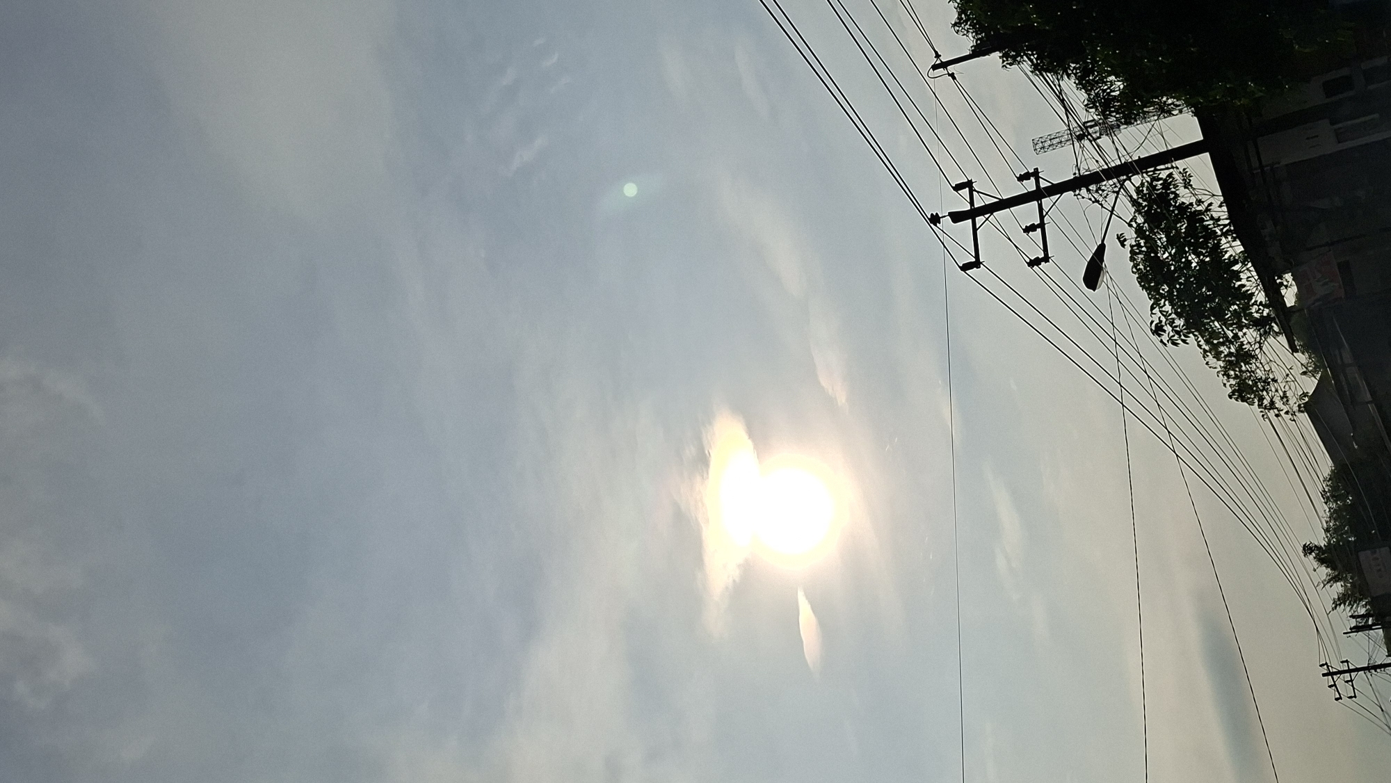 The image captures a view of the sky and a streetscape in Keudebayu, Indonesia. The sun is bright and partially obscured by clouds, creating a halo effect. Power lines crisscross the frame, supported by utility poles. A street lamp is visible on one of the poles, and trees and building silhouettes are present along the bottom of the image. The overall impression is of an overcast day with the sun attempting to break through.