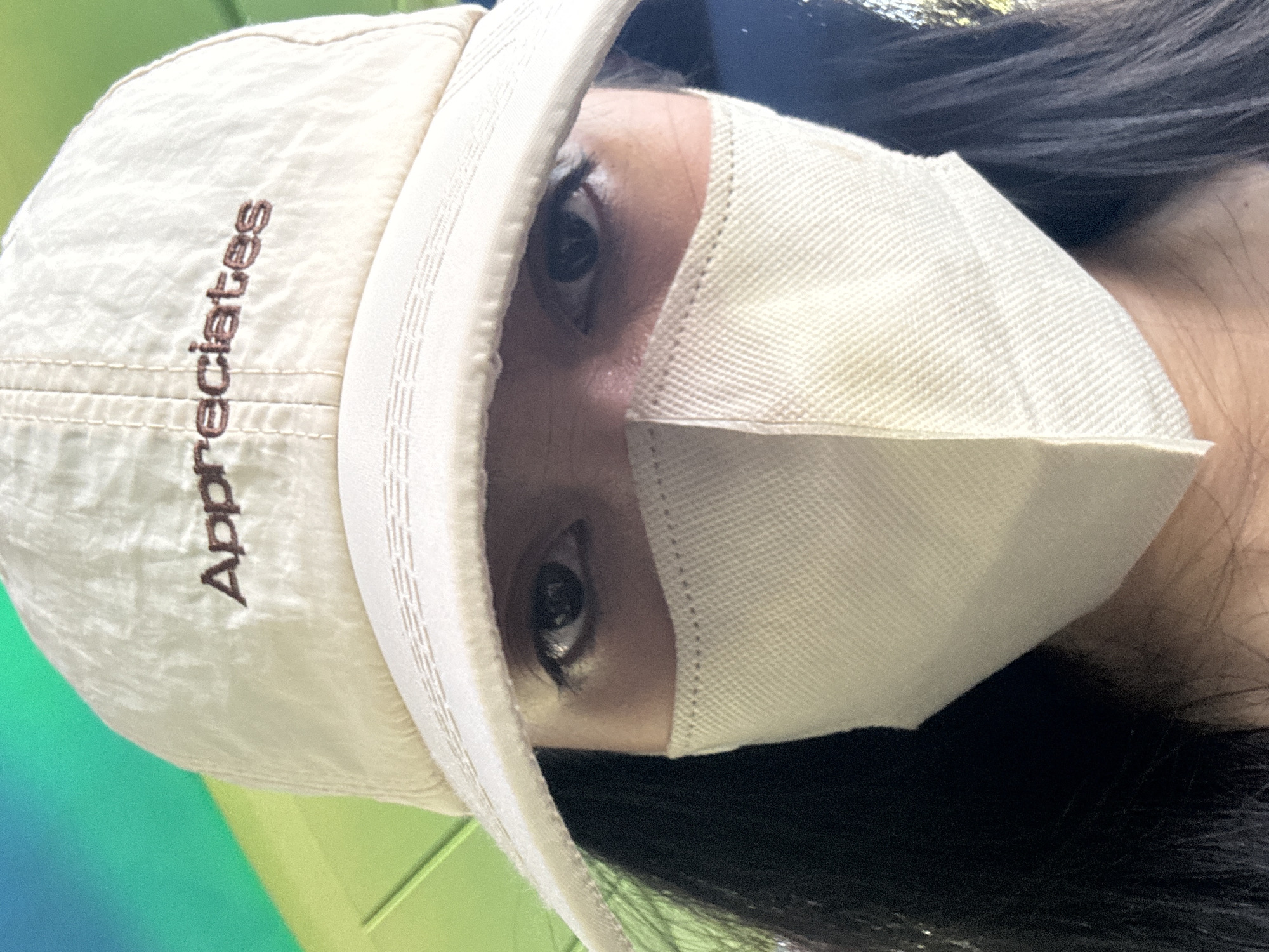 A close-up selfie shows a person's face from the eyes down, wearing a light-colored baseball cap and a light-colored disposable face mask. The cap has embroidered text in brown that reads "Appreciates." The background is out of focus but appears to be an outdoor setting with bright green and blue colors. The person has dark hair visible around the edges of the frame. The lighting suggests it is daytime. There is no indication of the location being Pudong, China.