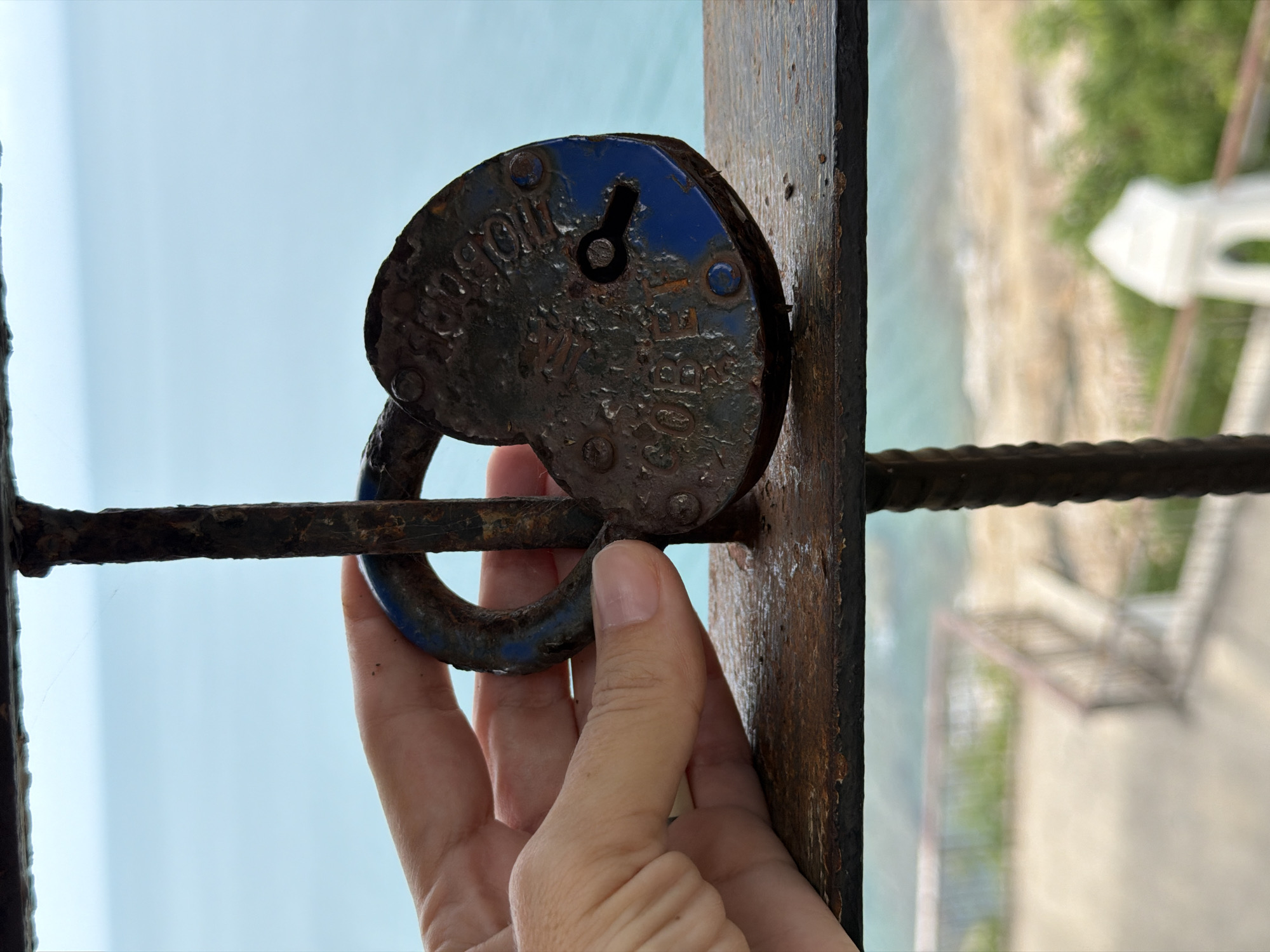 An aged, heart-shaped padlock, coated in rust and peeling blue paint, is being held by a person's hand against a rusted metal railing. The padlock has a visible keyhole. Behind the railing, the turquoise ocean stretches out under a pale sky, suggesting a coastal location. In the foreground, the railing appears to be part of a fence or structure overlooking a blurred background that includes vegetation and some white architectural elements. The overall scene suggests a viewpoint in a tropical setting, possibly a waterfront area in Ko Samui District, Thailand, where lovers' padlocks are sometimes left as symbols of commitment.