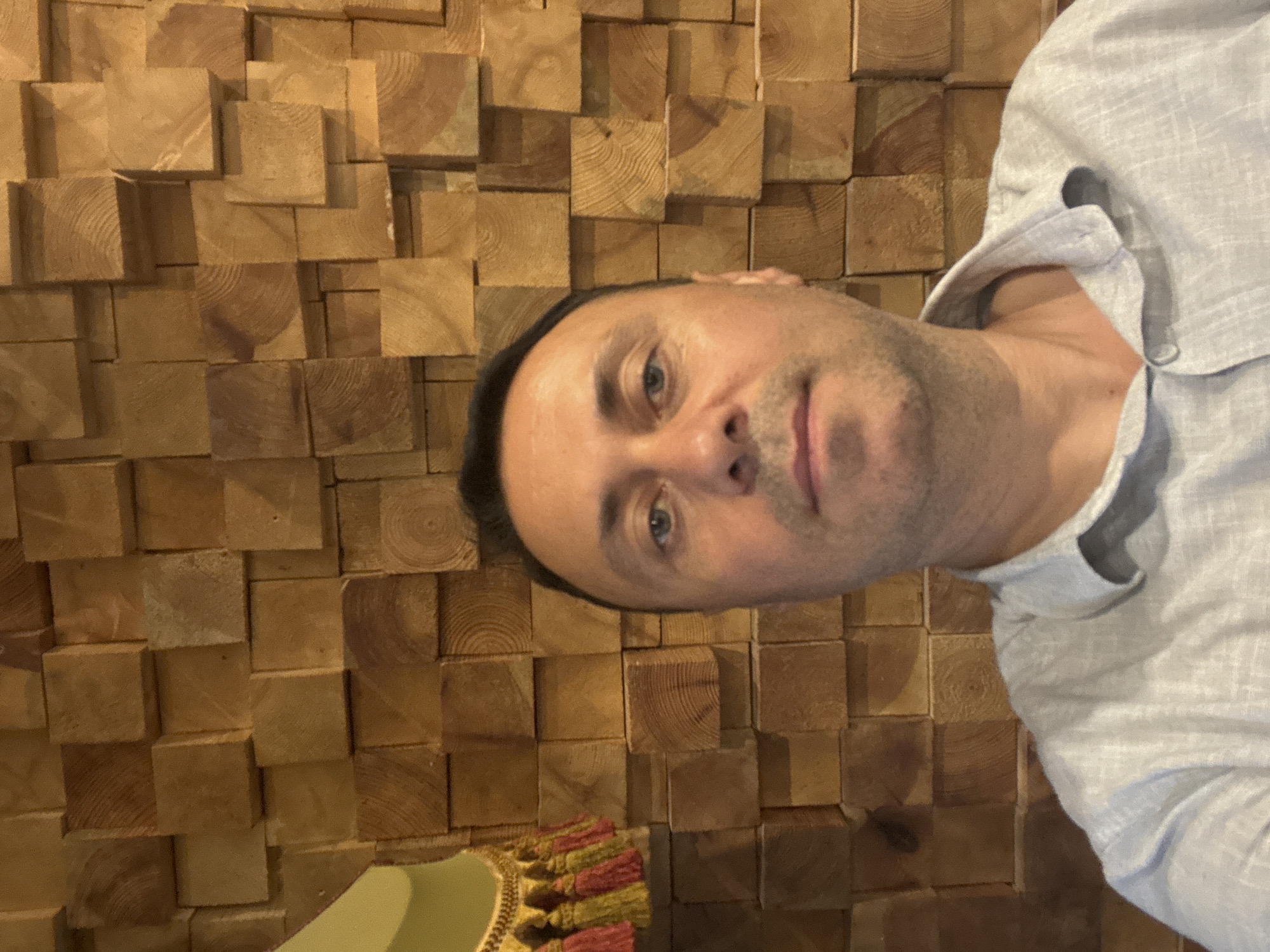 A man is taking a selfie in front of a wooden wall in Tbilisi, Georgia. The man is wearing a light blue collared shirt. The wall is made of wooden blocks of varying sizes and depths. To the left of the man, a decorative hanging with tassels is visible. The lighting is warm and appears to be artificial.