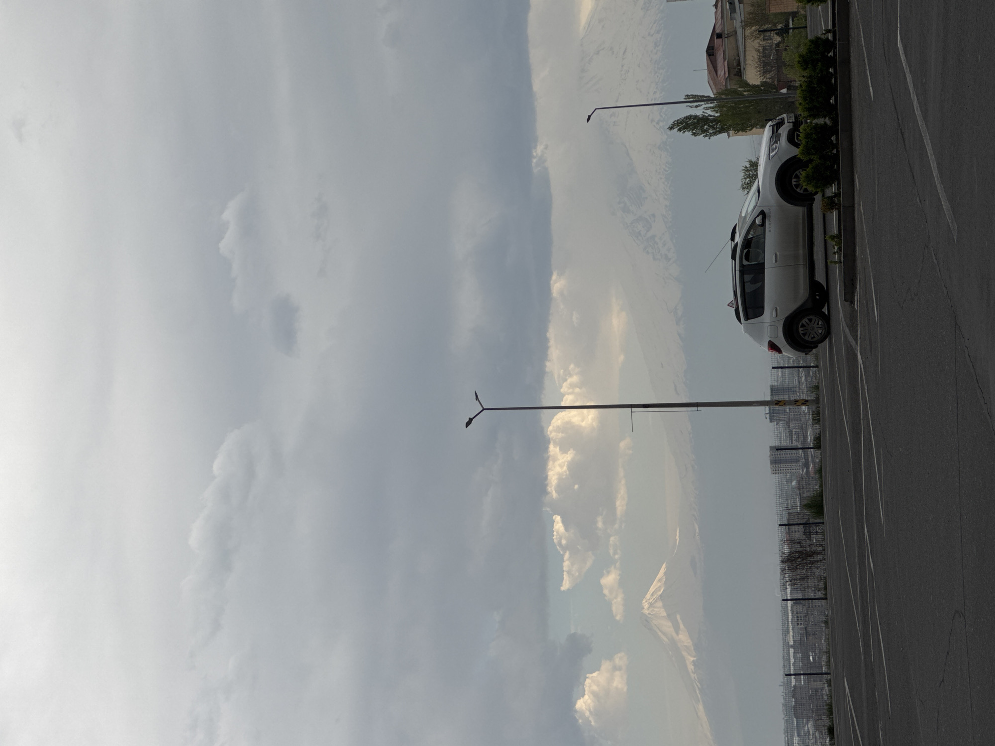 The image captures an expansive outdoor scene in Yerevan, Armenia, showcasing a blend of urban development and natural grandeur under a cloudy sky. In the right foreground, a white SUV or crossover vehicle, possibly a Dacia Duster, is parked on an asphalt surface marked with white lines, typical of a parking lot or roadside. Behind the car, a line of green bushes and small trees separates the paved area from various buildings further back.
A tall, dark streetlamp stands prominently on the left side of the frame, extending towards the sky. The middle ground reveals a broad cityscape with numerous buildings and structures, indicative of an urban environment. A metallic fence runs horizontally across this mid-ground, providing a visual boundary within the cityscape.
In the far background, the majestic, snow-capped peaks of Mount Ararat dominate the horizon. These iconic mountains are partially obscured by an extensive layer of grey and white clouds. The sky is largely overcast, creating a diffused light across the scene, suggesting daytime, possibly late afternoon, as some patches of brighter light break through the cloud cover. There are no people visible in the image, and no legible text can be discerned on any objects or structures. The overall mood is serene, emphasizing the harmonious juxtaposition of the city with its powerful natural backdrop.