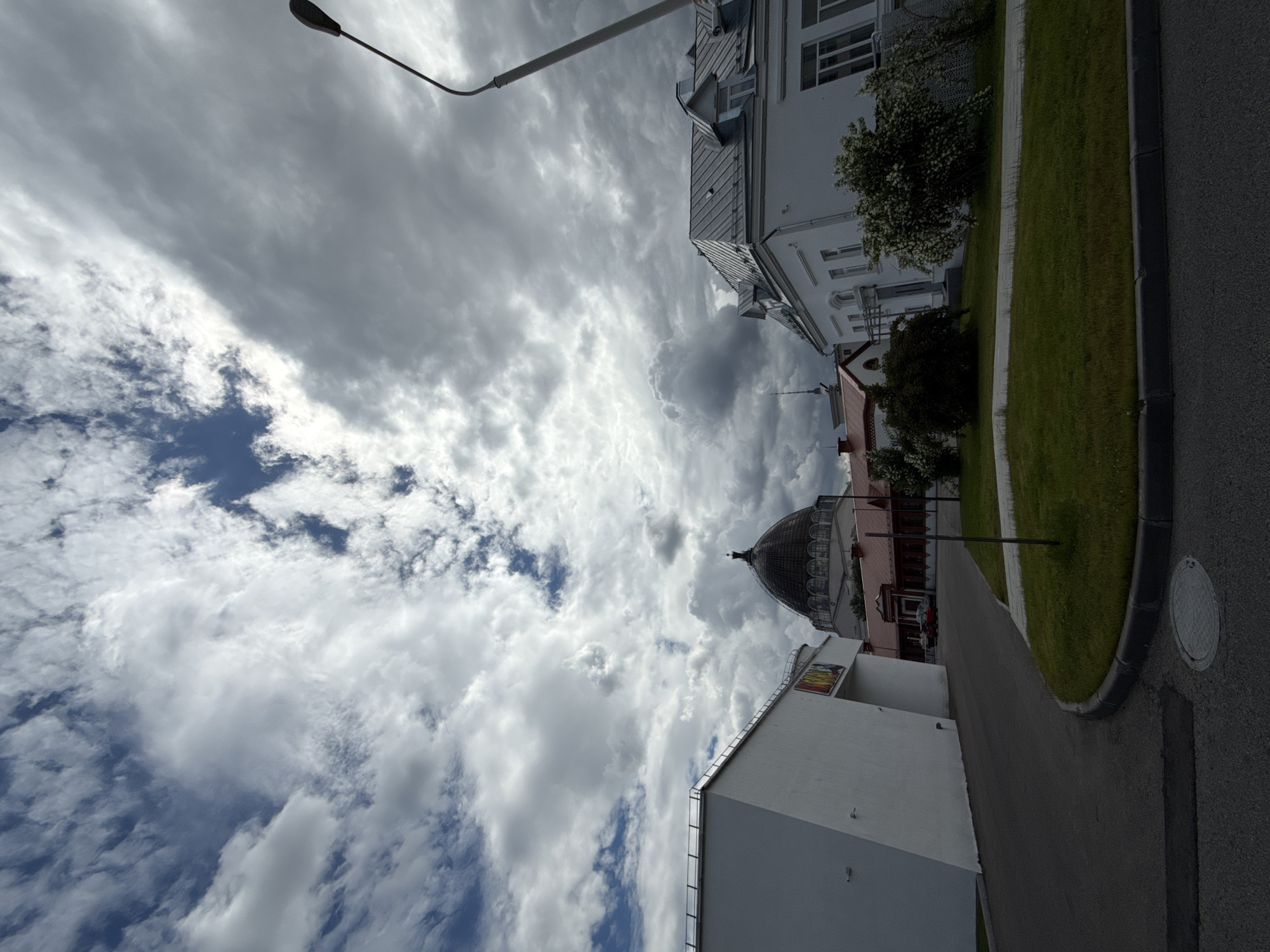The image depicts a street scene in Moscow, Russia, with a large, domed building in the background. The sky is filled with dramatic, dark clouds, suggesting an overcast or approaching storm. In the foreground, a paved road curves to the left, with a grassy area and a sidewalk bordering it. To the left of the road is a modern, white building with a unique geometric design. To the right, a white, two-story building with a metal roof and dormer windows is visible. The domed building in the center appears to be a significant architectural landmark, possibly a museum or government building, with a reddish-brown structure at its base. A tall, modern street lamp is positioned on the right side of the image, reaching into the cloudy sky. The scene appears to be devoid of people, with only a few cars visible in the distance near the domed building. The overall impression is one of an imposing and somewhat moody urban landscape under a turbulent sky.