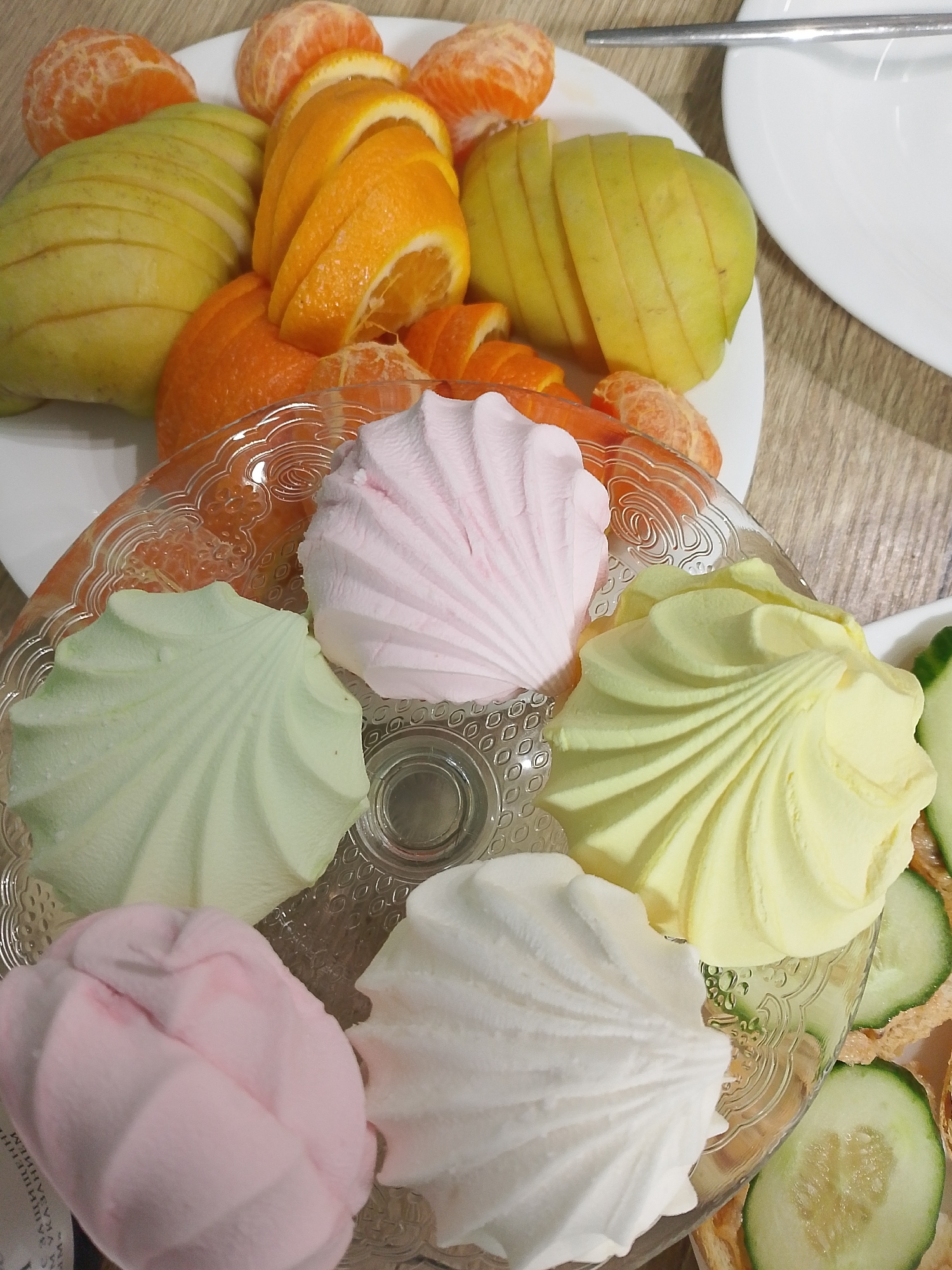 A high-angle, indoor shot depicts a glass serving dish containing five pastel-colored, seashell-shaped marshmallows. The marshmallows are arranged in a loose circle and are colored green, pink, yellow, pink, and white, respectively, going clockwise from the top left. Behind the serving dish, a white plate holds sliced fruit, including peeled tangerines, sliced oranges, and sliced pears or apples. A stainless steel utensil rests on the plate, and another white plate is partially visible to the right. The background appears to be a wooden table. Cucumber slices are located at the bottom right of the image. The image does not contain any identifying information that would allow determination of whether or not this image was captured in Izhevsk, Russia.