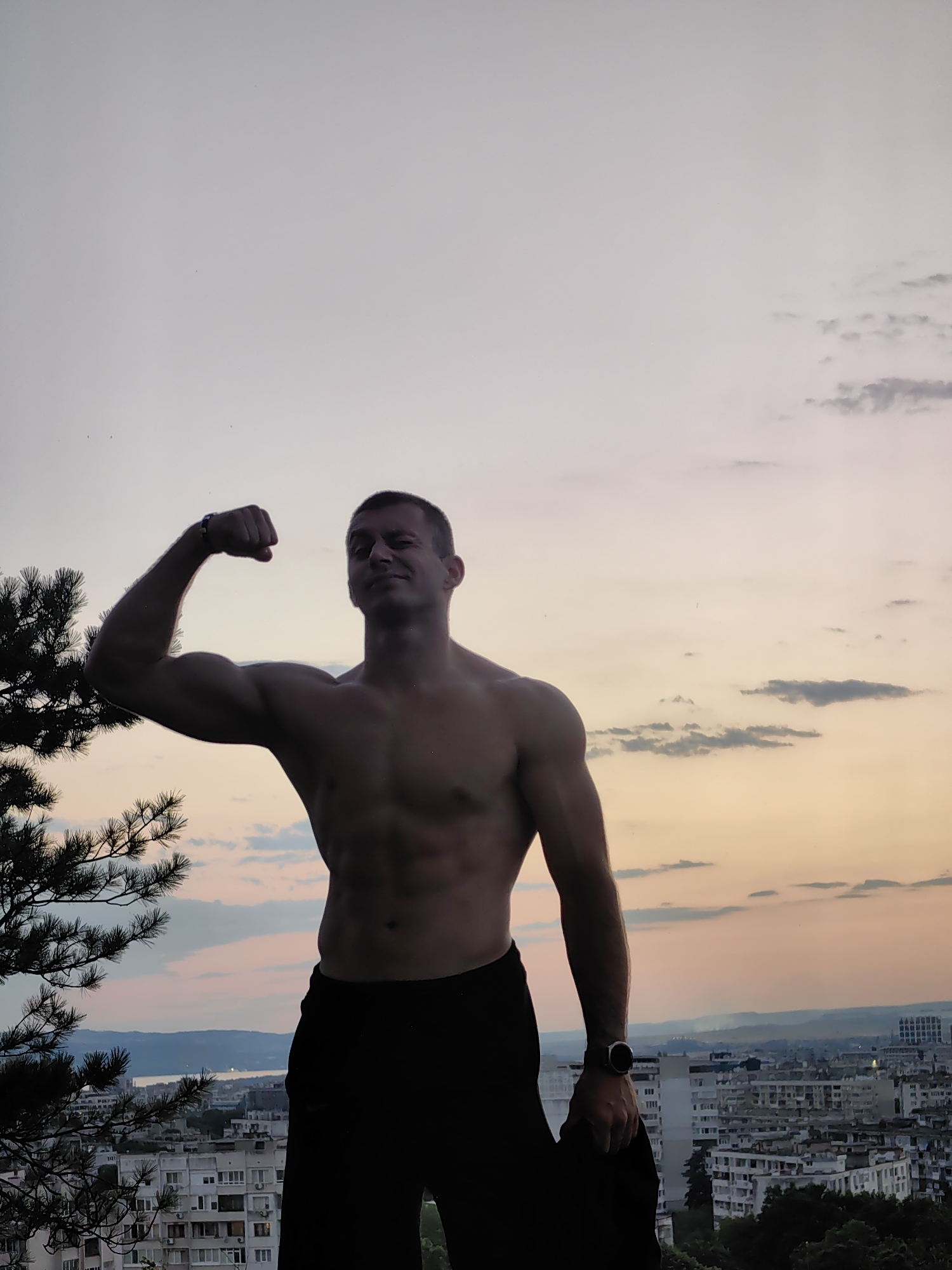 A shirtless man with defined abdominal muscles and biceps flexes his left arm, standing on a hill overlooking a city during sunset. He is wearing black pants and a smartwatch on his left wrist. A pine tree with dark green needles is positioned to the left of the man, partially obscuring the view of the cityscape. The sky is a gradient of soft orange, pink, and gray, with scattered clouds. In the distance, a body of water can be seen, with a city spread across the horizon, suggesting the location is Varna, Bulgaria, likely from a vantage point like the Asparuhovo bridge area or the surrounding hills. The buildings in the foreground are a mix of apartment blocks, some appearing aged. The lighting indicates it is either early morning or late evening.
