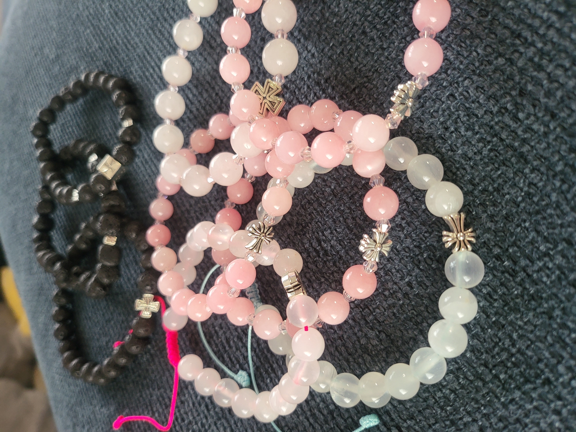 The image shows several beaded bracelets, some made of dark, matte beads and others of pink and white translucent beads. The bracelets are arranged on a dark, textured surface, possibly denim. Some of the bracelets are adorned with small, metallic charms shaped like crosses or flowers. There is a bright pink string attached to one of the dark bracelets, and a light blue string near the pink and white bracelets. The scene is indoors, with no indication of the city of Peski, Russia. There are no people or animals visible in the image.