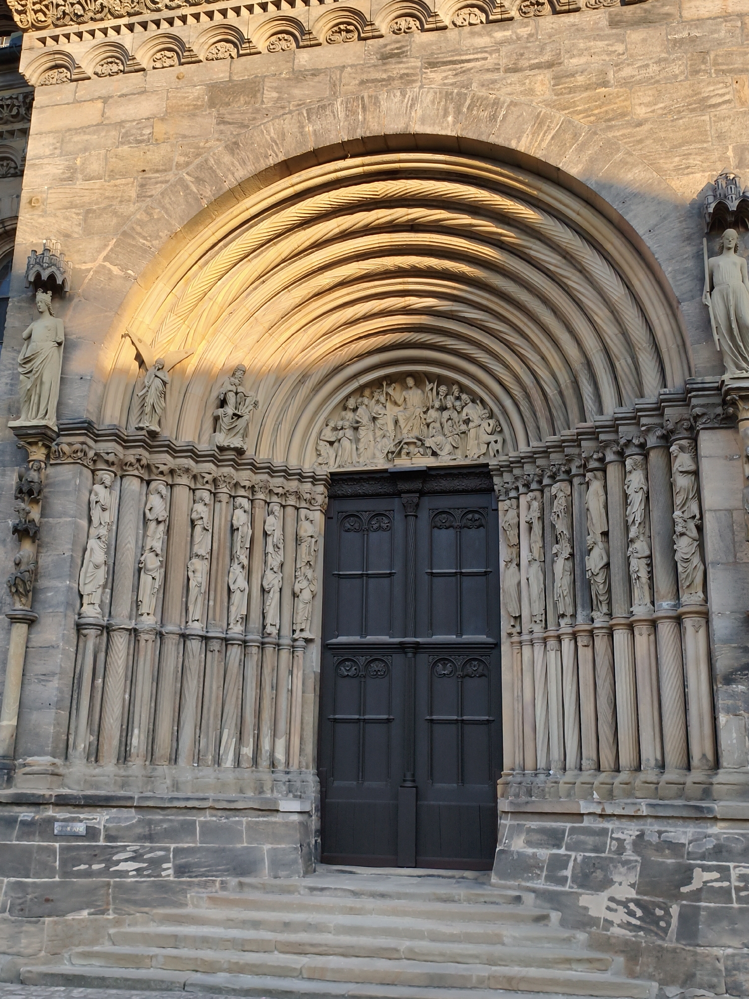 The image shows the west portal of Bamberg Cathedral, located in Bamberg, Germany. The portal is characterized by a large, semicircular archway constructed with deeply recessed, ornately carved concentric rings. These rings are further decorated with intricate patterns, creating a play of light and shadow. Below the arch, a dark wooden double door stands centrally. Flanking the door and archway is a colonnade of slender, decorative columns, each topped with a capital and supporting a horizontal frieze. Each column in the lower colonnade appears to be sculpted into a slender, standing figure, mostly draped in classical robes. Above the door and between the upper rings of the main arch, a relief carving depicting a biblical scene with multiple figures is visible. Stone steps lead up to the portal, and the building's facade is constructed from large, roughly hewn stone blocks. The warm, directional light suggests it is either early morning or late afternoon, casting long shadows across the stone surfaces.