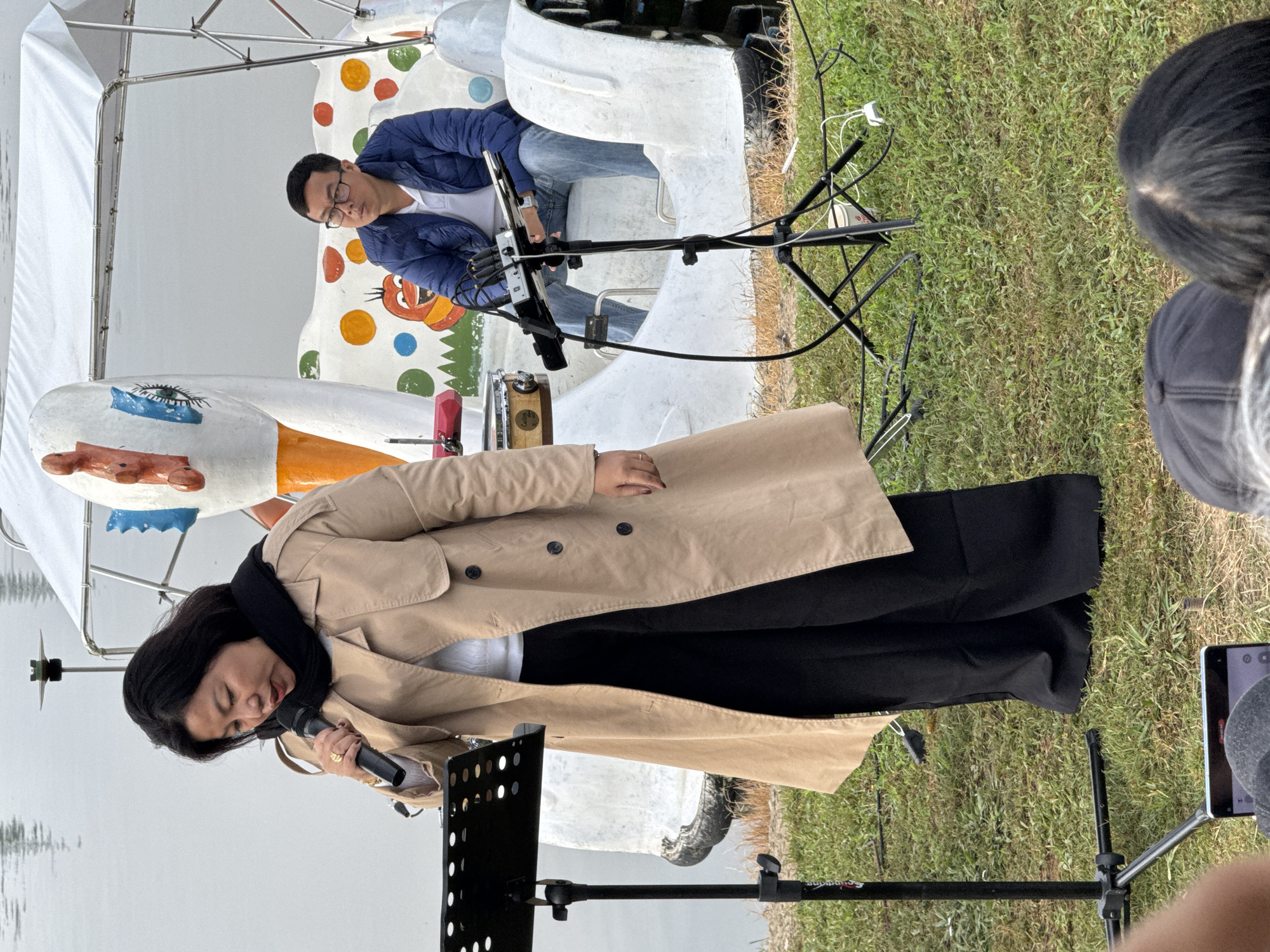 The image depicts an outdoor scene at the Xuan Huong Lake in Da Lat, Vietnam. In the foreground, a woman wearing a beige trench coat and black pants is speaking into a microphone, standing in front of a music stand. Behind her, a man in a blue puffer jacket and jeans sits on a boat decorated with a large swan design. The boat is white with colorful polka dots and a cartoonish orange creature. The background shows the calm waters of the lake and the grassy bank where the event is taking place. Several cables and a small drum are visible on the ground near the boat. The overall atmosphere suggests a casual outdoor performance or event.