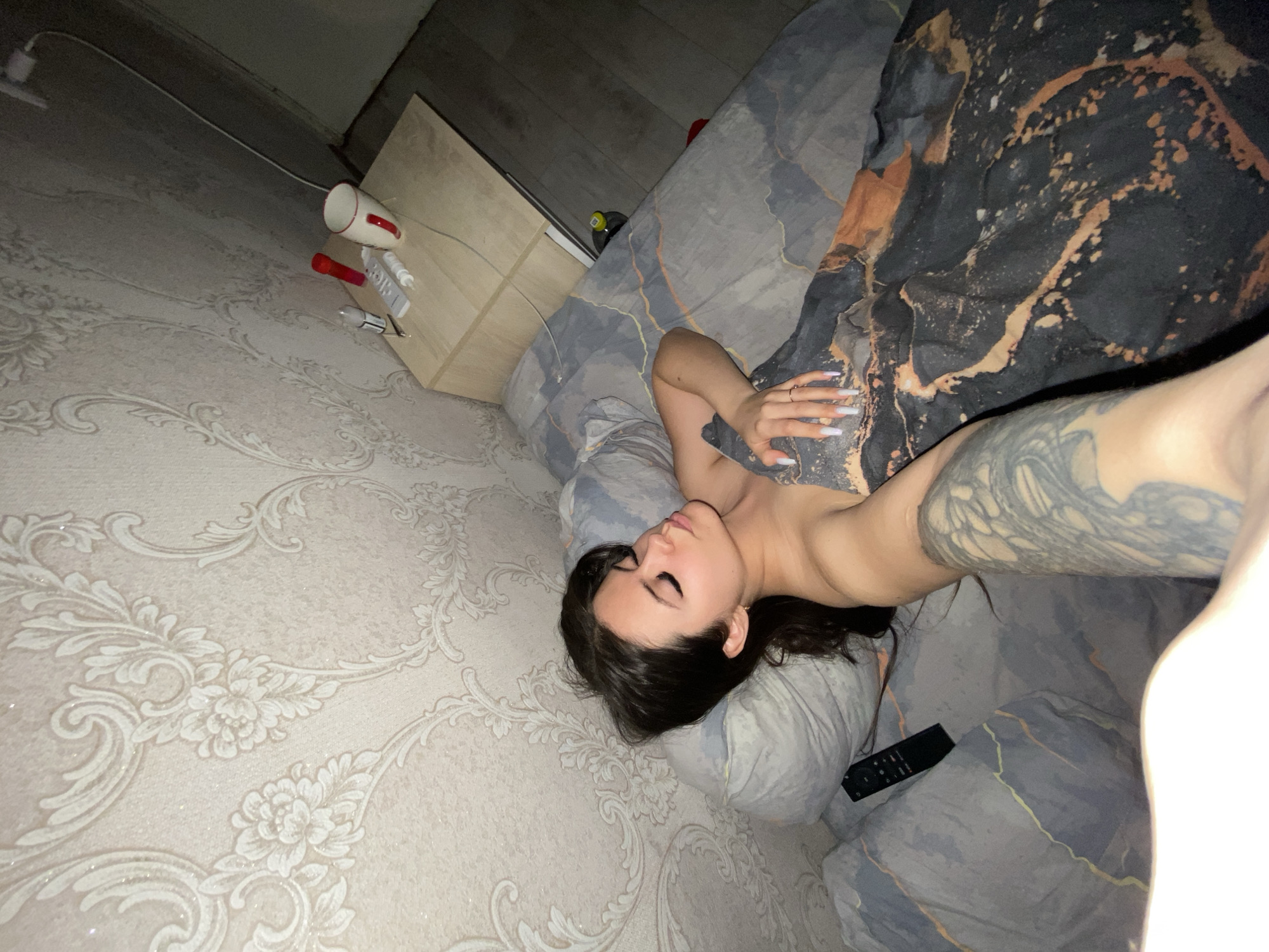 This image, captured in Chaikovsky, Russia, depicts a young woman resting or sleeping in a dimly lit bedroom. The perspective suggests a self-portrait, taken with a tilted camera angle. The woman, with dark hair, lies on her side with her eyes closed, showcasing prominent long, dark eyelashes. Her torso is bare, partially covered by a dark brown and black blanket featuring abstract light brown and orange patterns. Her right arm, which has extensive grey and black tattoos, is visible, extending towards the foreground. Her left hand, adorned with long, light-colored nails and a ring, rests on the blanket.
She is positioned on a grey pillow and a bed covered with grey bedding that has subtle abstract linear designs. A black remote control is visible on the bed near her head. Adjacent to the bed is a light wooden nightstand or small cabinet. On the nightstand, several objects are discernible: a white mug with a red handle, a red object (possibly a small flashlight or vibrator), a white remote control, and a cylindrical electronic device. A white power cable and adapter are also visible along the wall near the nightstand. The wall is decorated with light-colored wallpaper featuring an ornate floral and scroll pattern. In the upper background, a wooden or laminate floor is visible. The scene evokes a private and relaxed moment of rest, likely at night or in the early morning given the subdued lighting. No discernible text is present in the image.