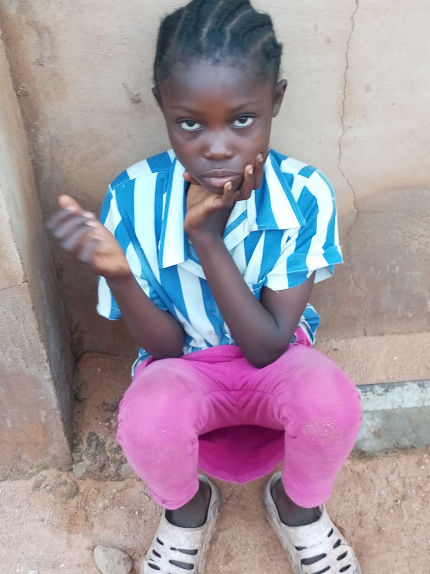 A young girl with dark skin and braided hair sits on the ground, leaning her head on her hand, and looking directly at the camera with a solemn expression. She wears a blue and white striped short-sleeved shirt and bright pink pants. Her feet are adorned with light-colored, perforated clogs. The background is a rough, light brown wall, and the ground beneath her is sandy with a few small stones. The lighting suggests it might be daytime, possibly late afternoon given the soft light and shadows. The scene appears to be an outdoor setting, likely in a rural or modest urban area, consistent with the location context of Tempure, Nigeria. There is no visible text in the image.