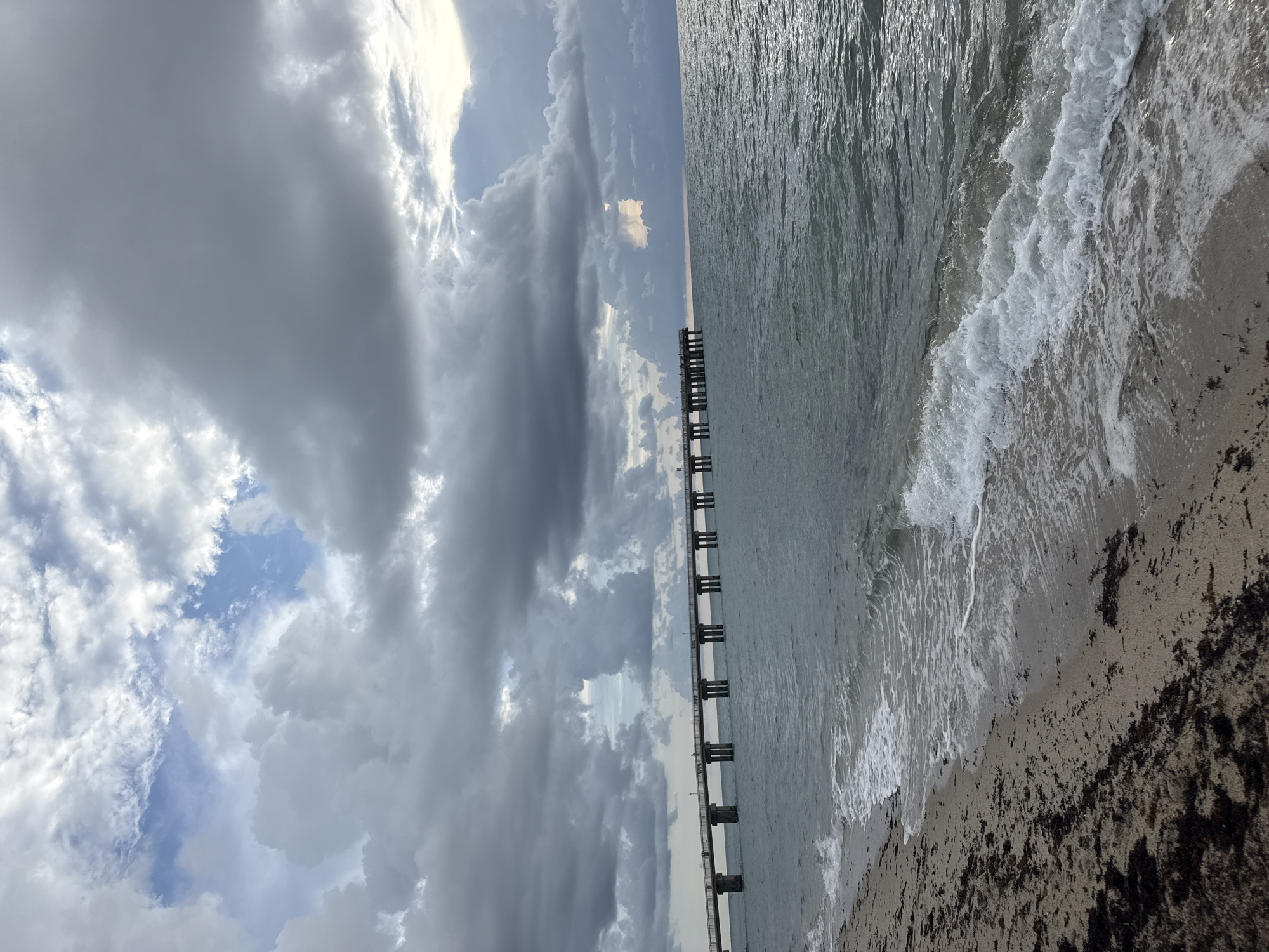 The image depicts a serene beach scene in Sunny Isles Beach, United States of America. The foreground features a sandy shore with scattered dark seaweed, leading to the gentle lapping of waves against the coast. A long pier, supported by concrete pillars, stretches across the horizon, with a few silhouetted figures visible on it. The ocean water is a muted blue-green, reflecting the dramatic, cloudy sky above. Large, grey clouds dominate the upper portion of the frame, with patches of blue sky peeking through in some areas. The overall atmosphere is calm and slightly overcast, suggesting a serene, perhaps pre-storm, moment.