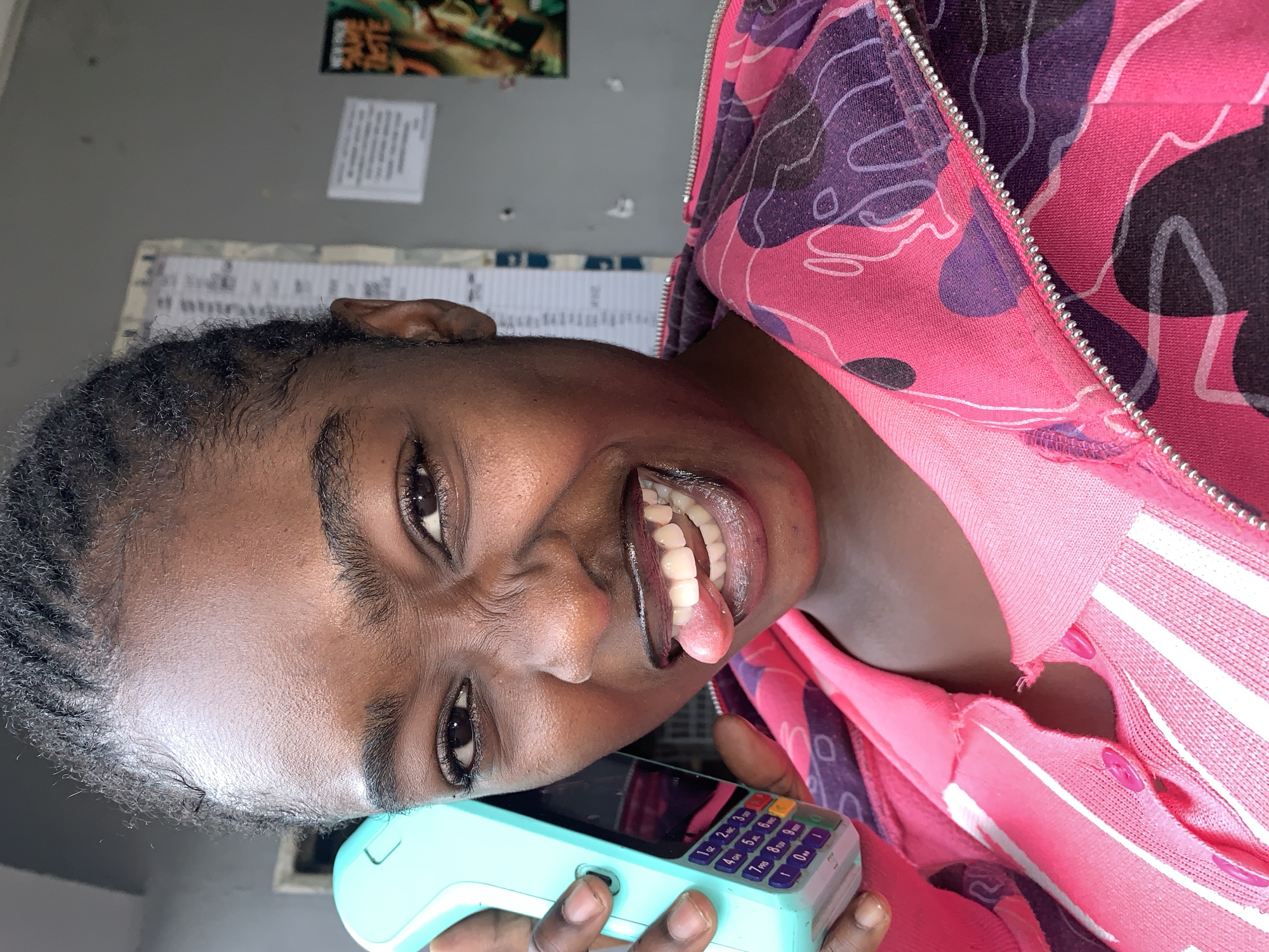 A close-up portrait captures a young Black woman in New Stadium, Nigeria, holding a turquoise point-of-sale device to her ear as if on a call, while sticking her tongue out playfully. She is wearing a vibrant pink zip-up hoodie with a purple and black abstract pattern over a pink ribbed top with white stripes. The background appears to be an indoor space, possibly a shop or office, with a bulletin board on the wall displaying a notice and a small poster with text that includes "NEW FADE TASTE." Her braided hair is pulled back, and she is looking directly at the camera with a wide, toothy grin.