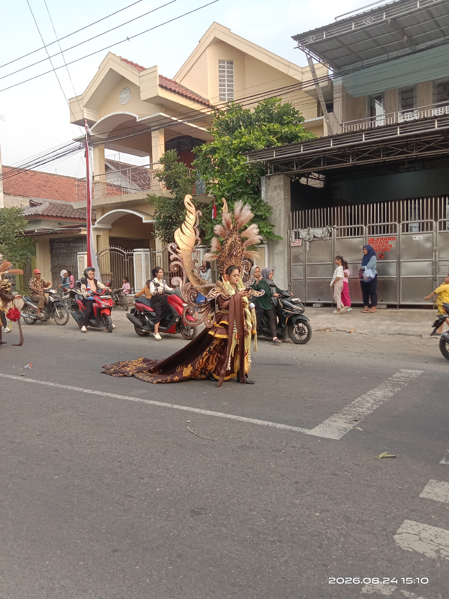 In Gudo, Indonesia, on August 24, 2026, at 15:10, a parade is in progress on a paved street. A person in elaborate traditional costume, featuring large, golden, feathered headdress and a long, patterned brown skirt, is the central figure. The individual is walking on the road, with their right hand holding a mobile phone. Behind them, a woman on a red motorcycle wears a light pink headscarf and a light pink top. To the left of the central figure, another woman on a dark green motorcycle wears a dark green headscarf and a dark green jacket. Further left, a man wearing a brown patterned shirt and a red helmet rides a motorcycle. In the background, two houses are visible. The house on the left has red tile roofing and a beige facade. The house on the right features a metallic gate and a beige facade with horizontal window blinds. Several other individuals are present, some standing and watching, others on motorcycles. A red and white flag is positioned on a pole to the left of the house. The sky is clear and appears to be late afternoon.