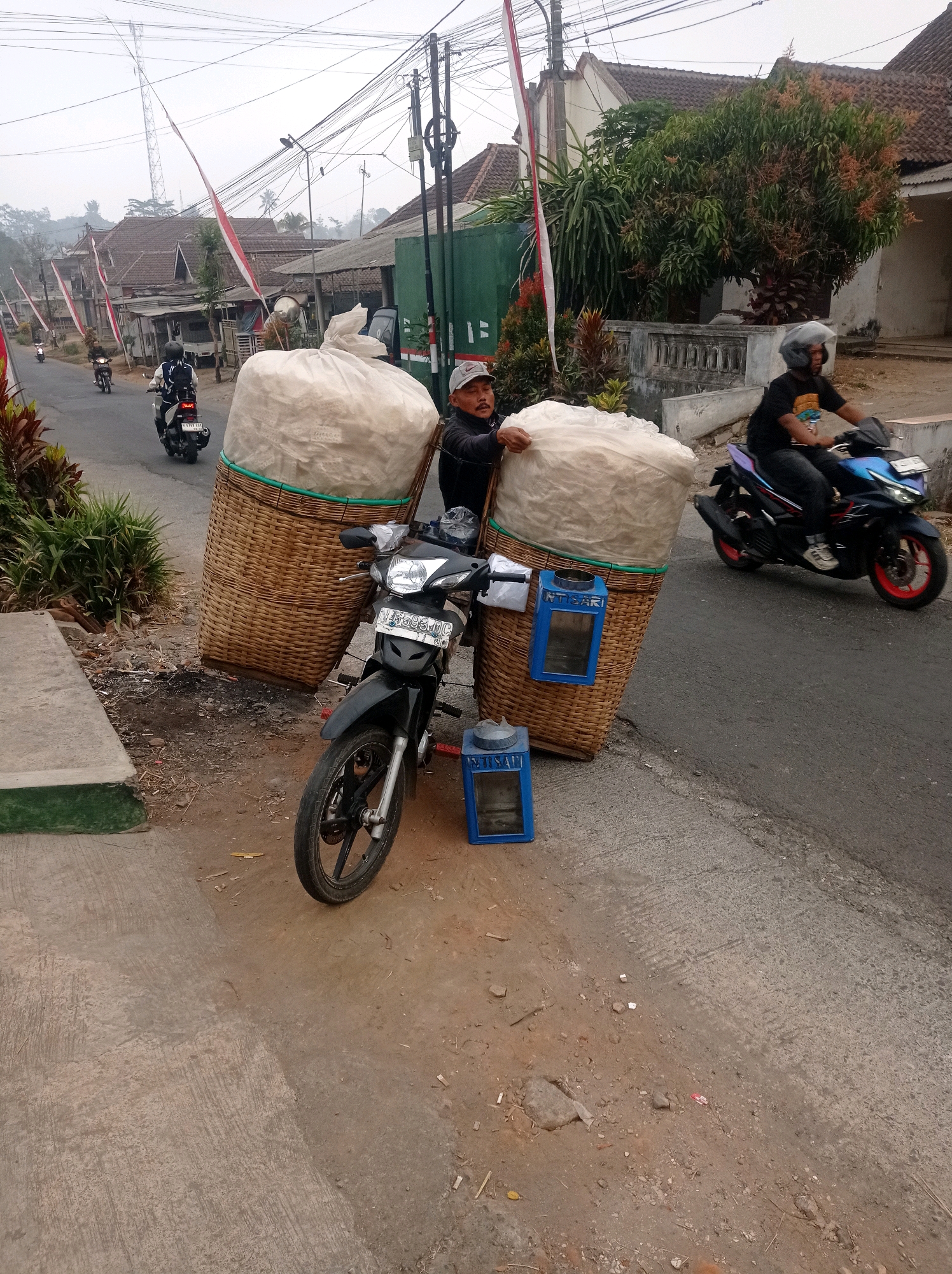 The image depicts a street scene in Purwosekar, Indonesia, with a man standing next to a motorcycle loaded with large woven baskets filled with light-colored material, possibly snacks or food items. The man, wearing a cap and dark clothing, is holding onto one of the baskets. A motorcycle with a license plate is parked in front of the baskets. Two blue containers, possibly for payment or trash, are attached to the baskets. Another person is riding a motorcycle in the background, moving away from the viewer. The environment appears to be a rural or semi-urban area with houses, trees, and utility poles visible. Indonesian flags are also present, suggesting a local event or national pride. The overall atmosphere is that of a typical day in a Indonesian town, with commerce and daily life in motion.