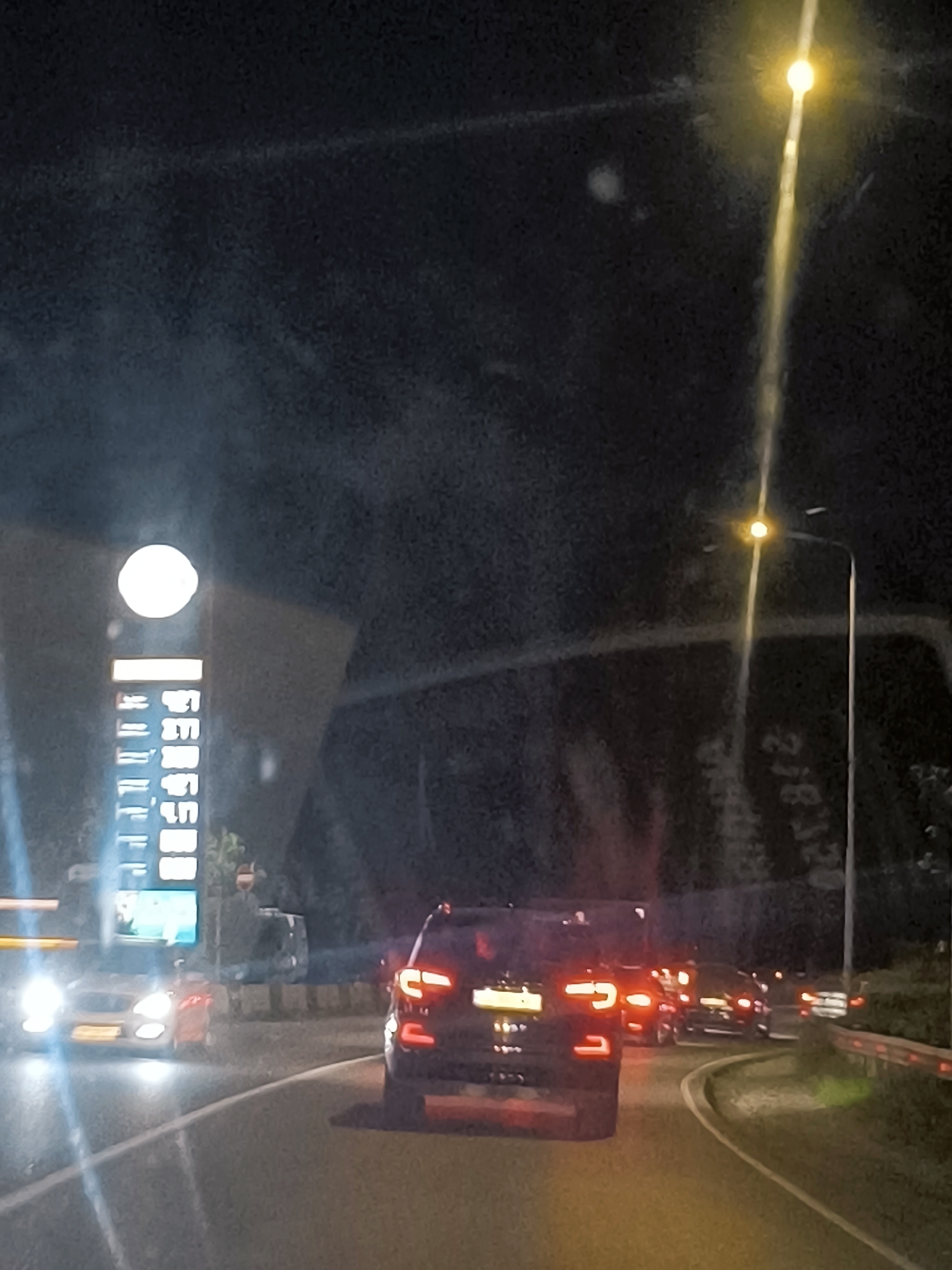 The image depicts a nighttime scene on a road in Batumi, Georgia, likely an urban or suburban area given the presence of streetlights and a tall illuminated sign. The perspective suggests the photo was taken from inside a vehicle. Several cars are visible, suggesting traffic. In the foreground, a dark-colored SUV is prominently displayed, its taillights illuminated. To the left, a white car with its headlights on is partially visible, and further back, other vehicles are seen with their lights on. A large, vertical, illuminated sign with rows of text and a circular light at the top stands to the left of the road, possibly indicating a business or service, such as a gas station. A tall streetlamp with a bright light source illuminates the right side of the image. The road surface is dark asphalt, and there are visible lane markings. The background is dark, with some faint outlines of structures or trees. There are no people clearly visible.