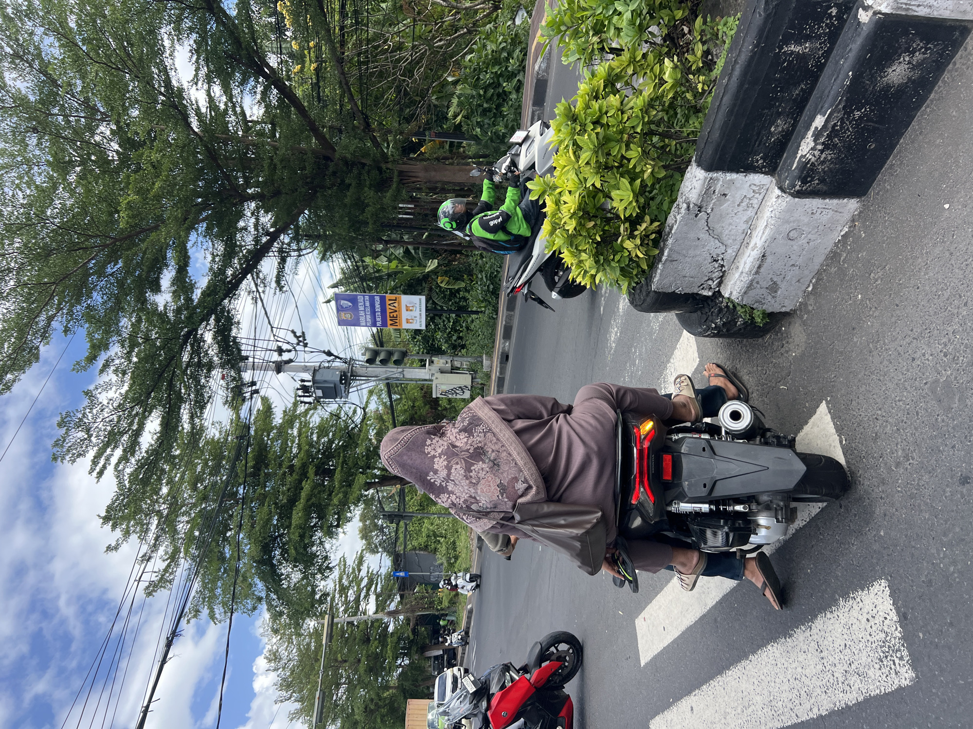 This image shows a street scene in Denpasar, Indonesia, featuring two motorcycles. In the foreground, a woman wearing a hijab and a long, brown dress rides as a passenger on a black motorcycle. She has a brown bag slung over her shoulder and is wearing flip-flops. The motorcycle is stopped at a pedestrian crossing. In the mid-ground, a person wearing a bright green jacket and a helmet, with a backpack, rides a white motorcycle. This individual appears to be a delivery driver, indicated by the logo on their jacket. They are riding on the right side of the road. The background shows lush green trees, utility poles with numerous wires, and traffic lights. A sign with Indonesian text and a police emblem is visible on the left side of the road. The overall environment suggests a busy urban intersection.