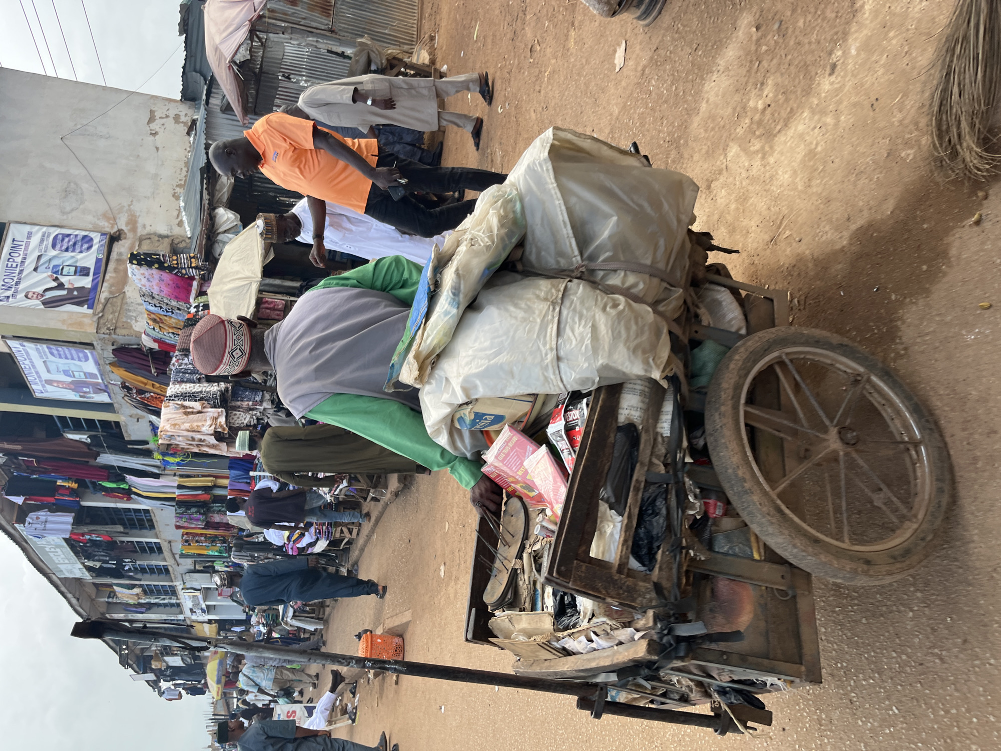 This image depicts a bustling market scene in Bukuru, Nigeria. In the foreground, a man wearing a green long-sleeved shirt and a flat cap is pushing a heavily laden cart. The cart, made of wood and metal, contains various items, including what appear to be books or pamphlets, plastic bags, and other unidentifiable objects. A sandal with nails protruding from it is prominently displayed on top of the contents.
Behind the man with the cart, several other individuals are visible. To the left, a man in a grey robe and flat cap walks away from the camera. Further back, a man in an orange polo shirt and black trousers is looking towards the right, holding a mobile phone. Other people, some carrying bags or wearing traditional attire, are scattered throughout the scene, suggesting active commerce.
The environment is an outdoor market with stalls lining the street. The stalls are adorned with clothing and other goods. The ground is unpaved and dusty, typical of a market area. In the background, advertisements are displayed on the buildings, one of which is for "MONIEPOINT," an electronic financial service provider. Umbrellas are set up in front of some stalls, likely for shade. The overall atmosphere is one of lively activity and commerce within an urban market setting.