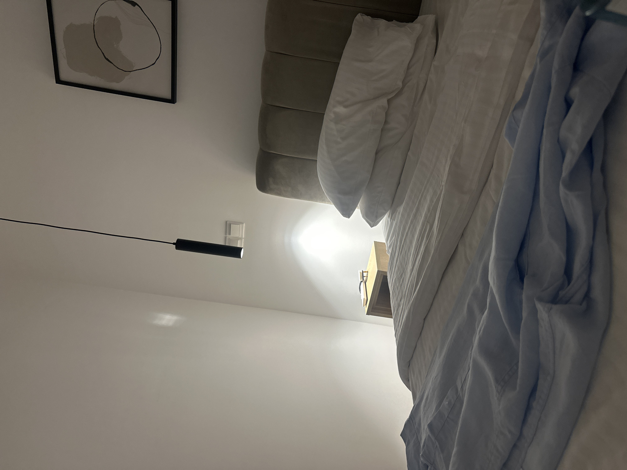 The image depicts a dimly lit bedroom interior, likely in a hotel or residential setting, in Larnaka, Cyprus. The scene is dominated by a large bed with white linens and two white pillows stacked on top. A light blue crumpled shirt lies on the left side of the bed. To the right of the bed, a small wooden nightstand is visible, upon which rests a pair of eyeglasses and a book. Above the nightstand, a wall-mounted light fixture casts a warm glow. Further up the wall, a framed abstract artwork hangs, featuring organic shapes in muted tones. A modern black pendant light fixture hangs from the ceiling, casting a beam of light onto the wall. The walls are painted a neutral, light color, and the overall ambiance suggests a quiet, perhaps evening, setting. There are no people visible in the frame.