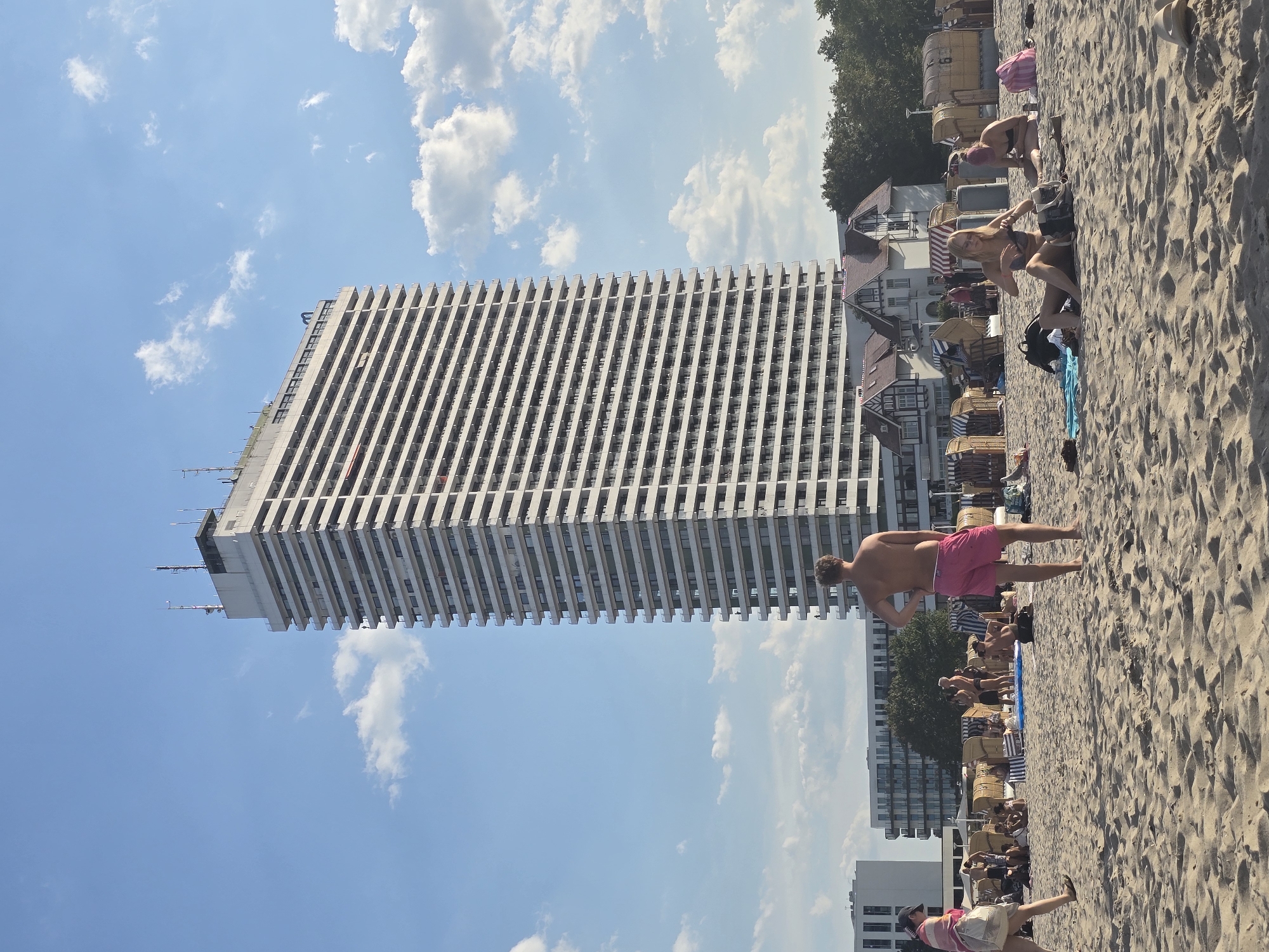 This is a full, vertical shot of a beach in Travemünde, a seaside resort area of Lübeck, Germany, on a sunny day. In the background, a tall, white high-rise building with many balconies dominates the skyline. In front of the high-rise, there are several smaller, traditional-looking buildings with pitched roofs, some with ornate gables. Trees line the edge of the beach area behind these buildings.
The beach itself is sandy, with many beach chairs, some covered with blue and white striped fabric, and others made of wicker. People are scattered across the beach, some sitting in the chairs, others lying on towels, and some are standing or walking.
In the foreground, a man with short, blond hair and wearing pink swim trunks stands facing away from the camera, with his hands on his hips. To the left of him, a woman with dark hair is walking away from the camera, wearing a striped cover-up over her shoulders and carrying a large tote bag. In the middle ground, a group of people are visible, some standing and talking, others sitting. To the right, two women are sitting on the sand in swimsuits. One is adjusting her bikini top, and the other, with pink hair, is looking to her right. Other beachgoers and objects like towels and bags are also visible throughout the scene. The sky is blue with scattered white clouds.
