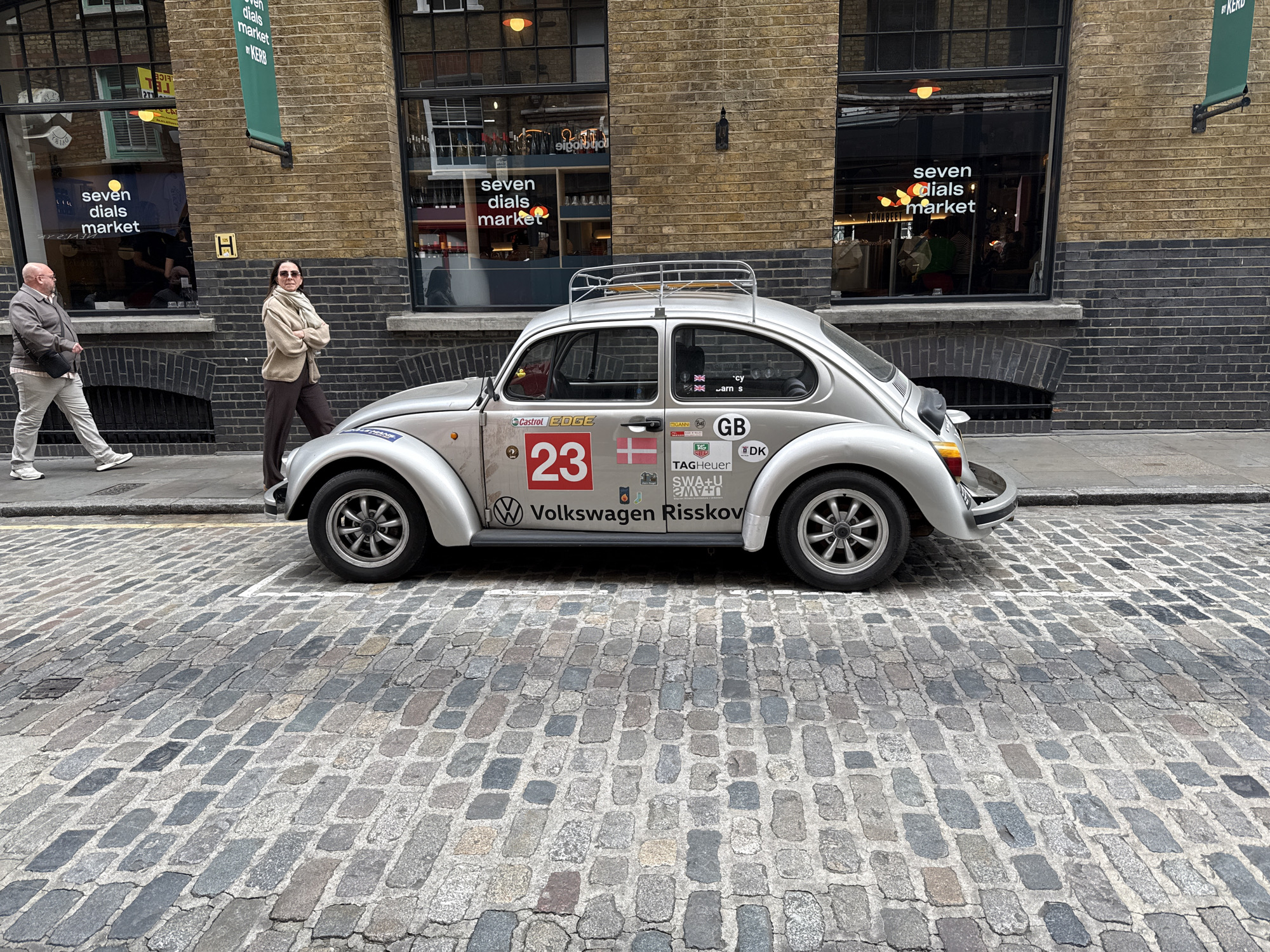 The image depicts a silver Volkswagen Beetle parked on a cobblestone street next to a building in London, United Kingdom. The building is made of brick and has two large windows with the words "seven dials market" displayed on them. A green banner with the words "seven dials market by KERB" is also visible on the building. A man in a tan jacket and grey pants is walking in the background, and a woman in a beige jacket and brown pants is standing with her arms crossed in front of the car. The car is decorated with stickers and decals, including the number "23" and the words "Volkswagen Risskov". The car also has a roof rack.