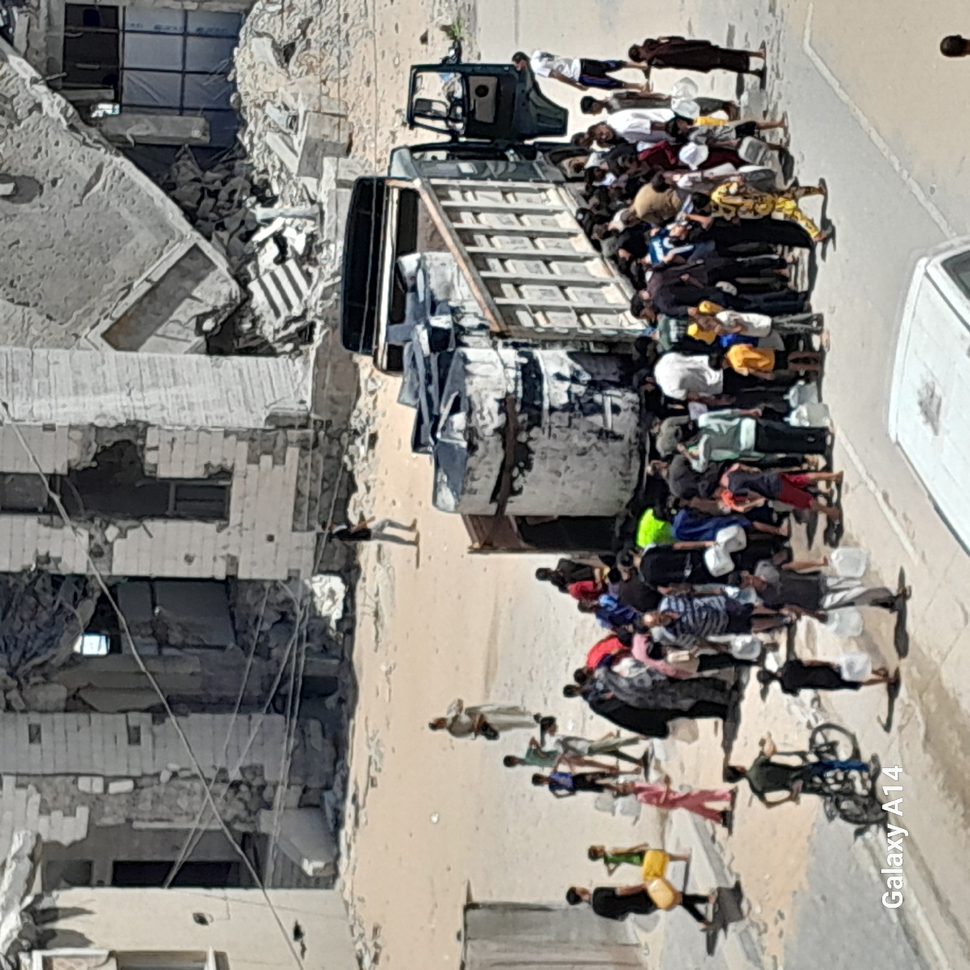 In Deir el-Balah, Palestinian Territories, a group of people are gathered around a damaged truck loaded with large water tanks. The truck is positioned on a dusty street next to partially destroyed buildings, some of which show significant structural damage. Many individuals, including children and adults, are carrying empty or filled water containers, suggesting they are collecting water. Some people are standing in line, while others are interacting with the truck or conversing amongst themselves. The overall environment appears to be one of scarcity and humanitarian effort, likely in a conflict-affected area.